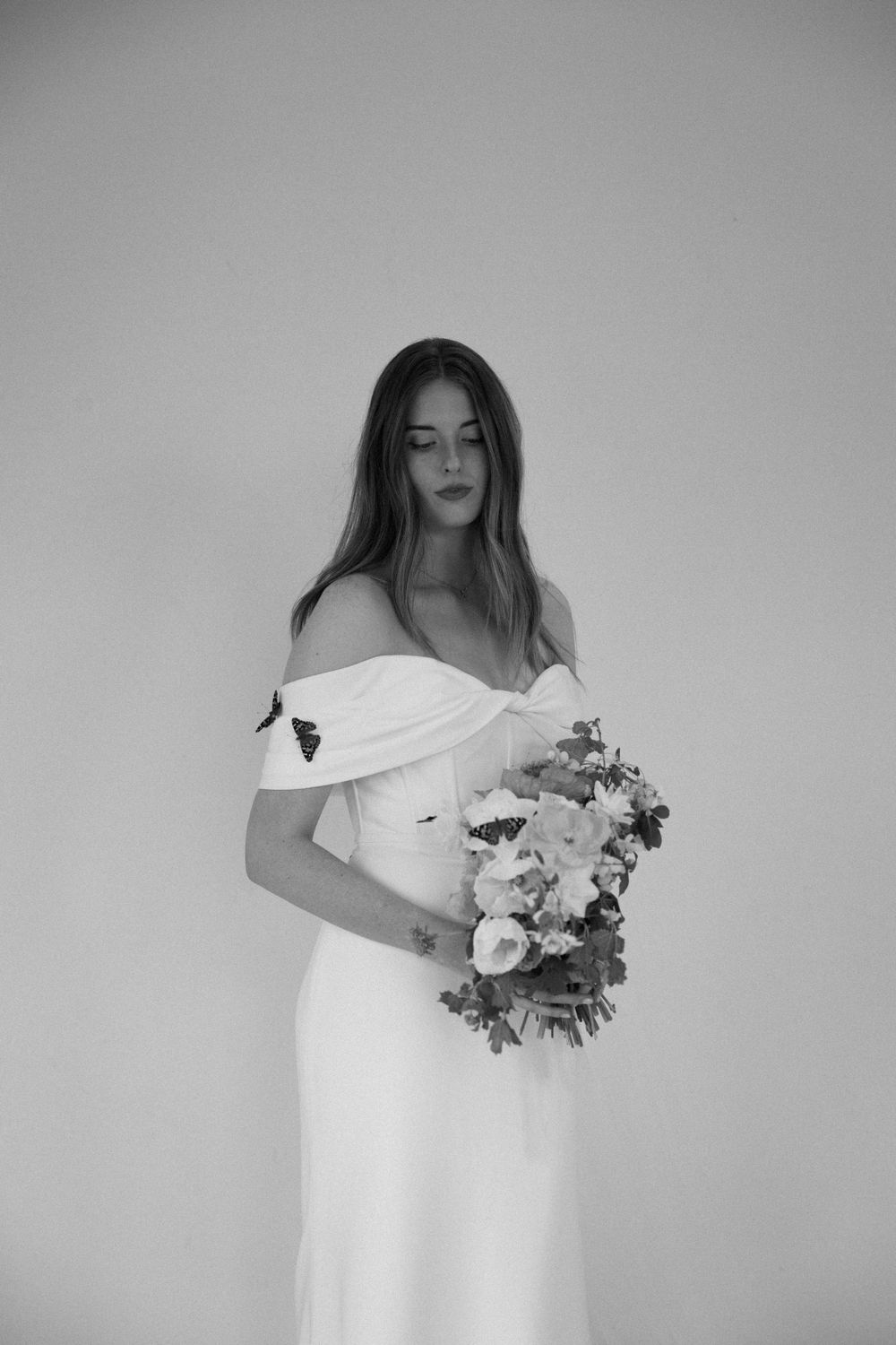 Black and white photo of a bride in a classic off-shoulder gown holding a lush floral bouquet against a plain backdrop.
