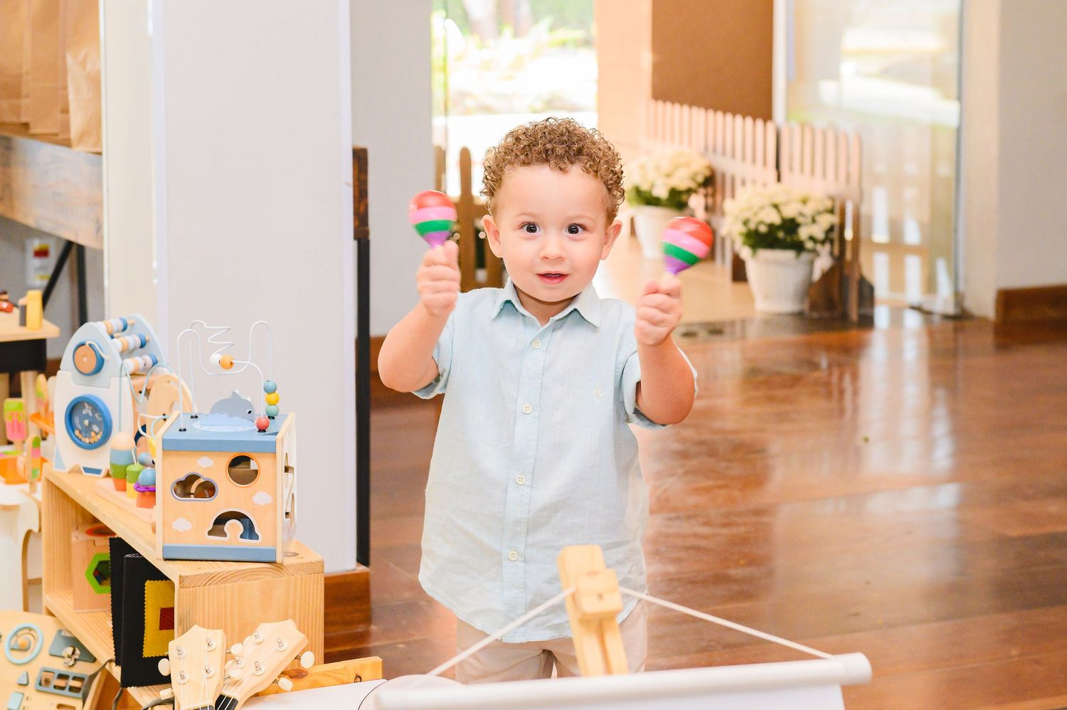 A young child wearing a light blue shirt plays with toys while standing at a wooden table in a bright indoor setting.