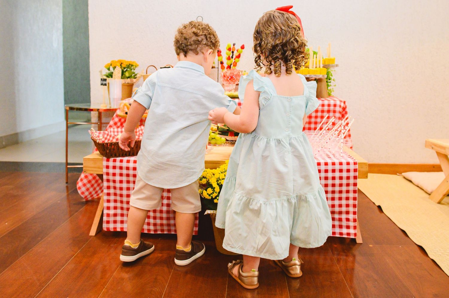 Two children in white outfits stand at a table with red checkered tablecloths and yellow flowers at a party setting.