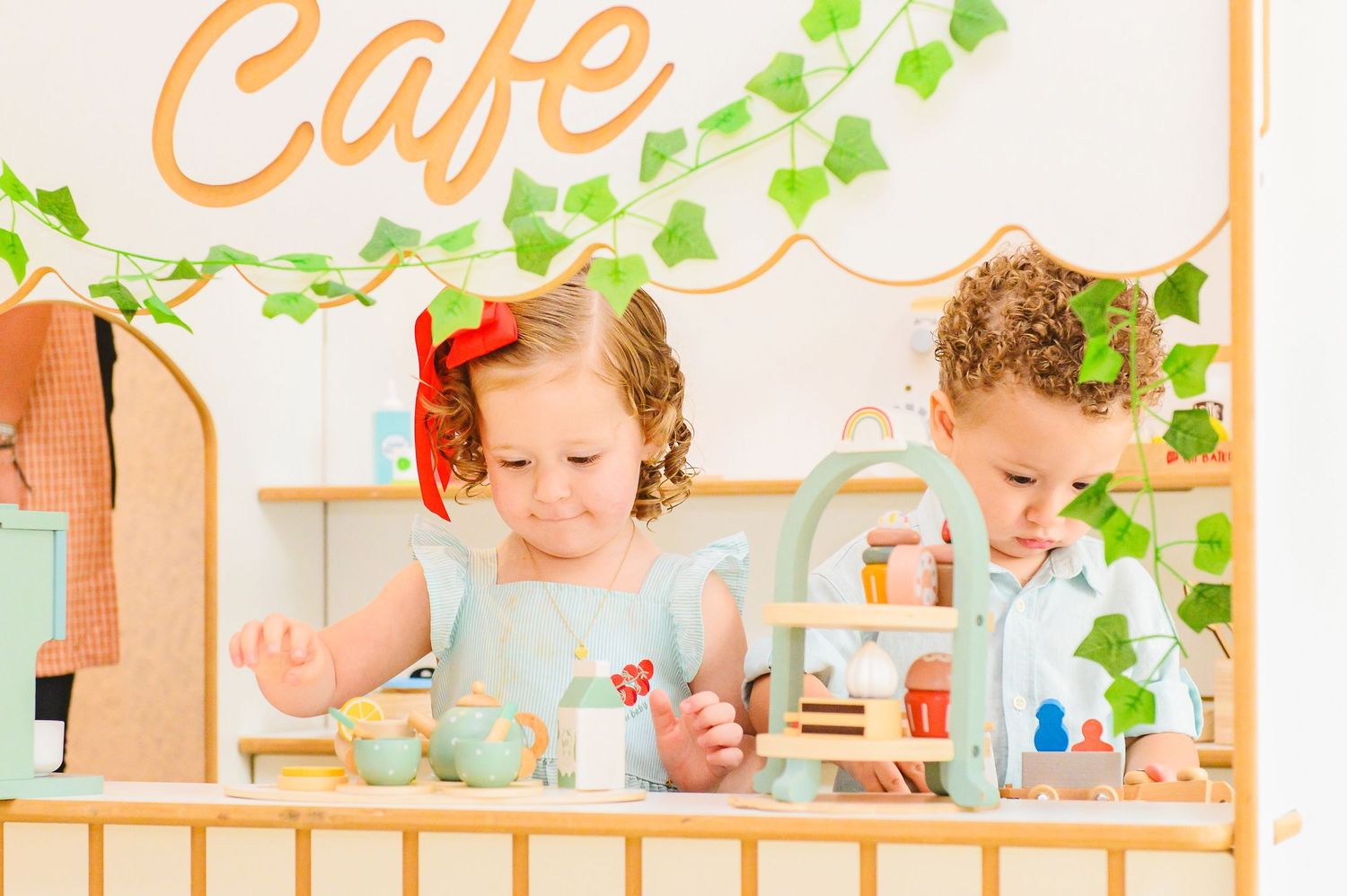 Two children play together at a pretend cafe with a wooden storefront and toy kitchen accessories while supervised.
