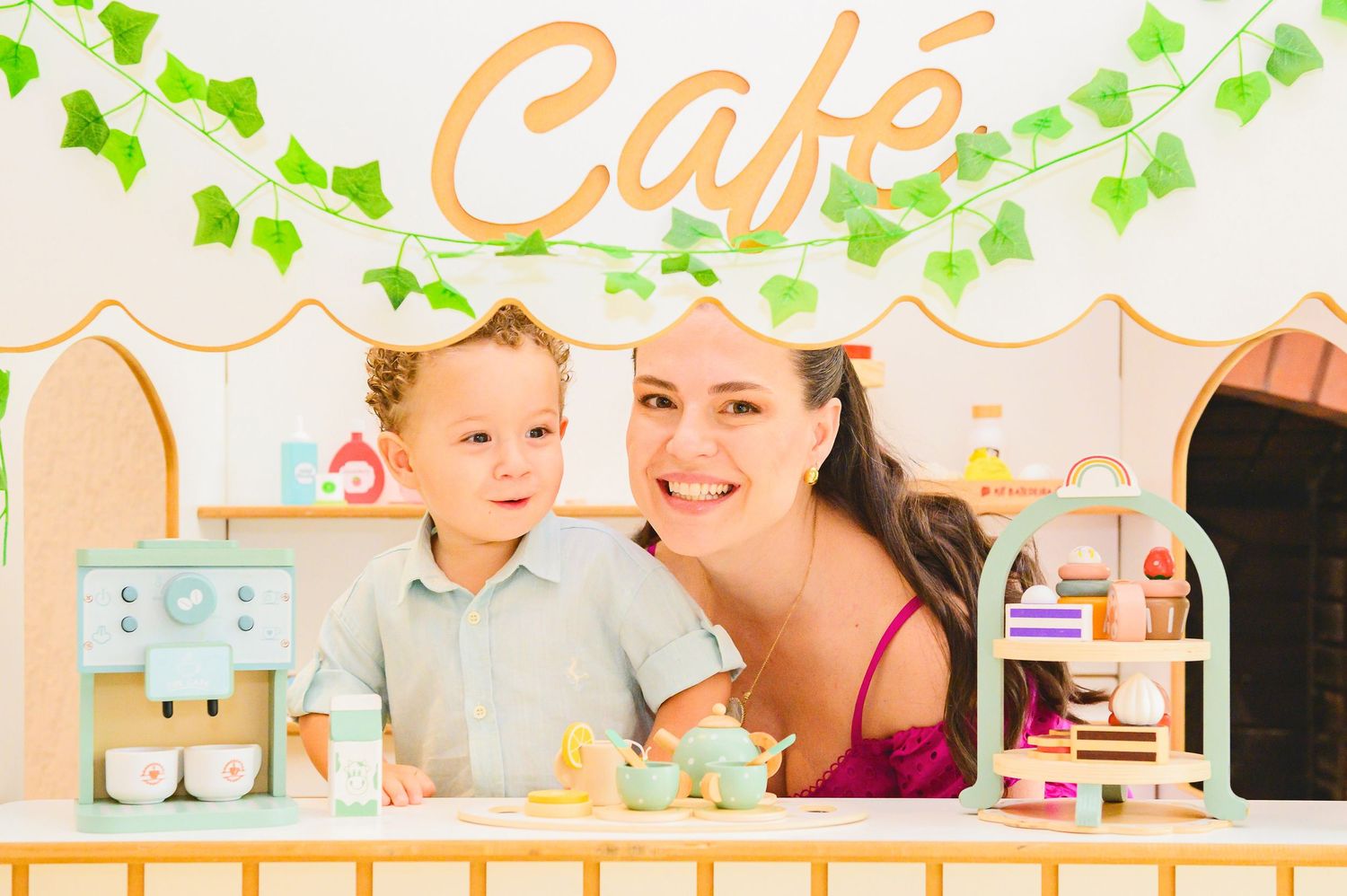A child and caregiver play together at a toy cafe counter decorated with green vines and peach-colored lettering.