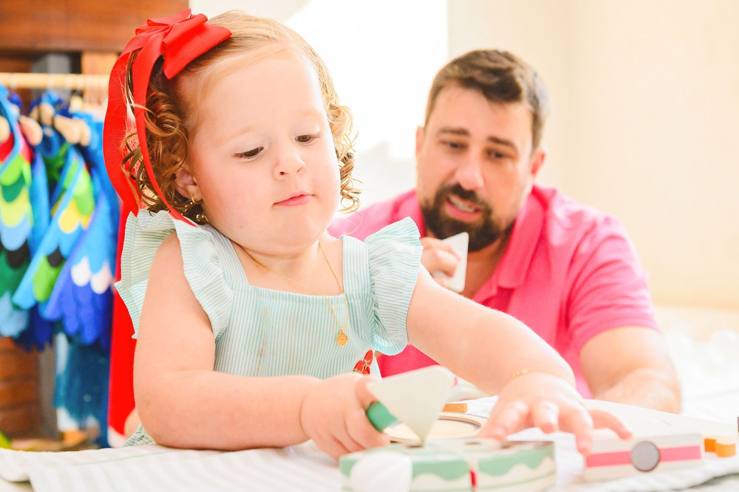A young child in a light blue outfit and red bow plays with art supplies at a table while a caregiver looks on.