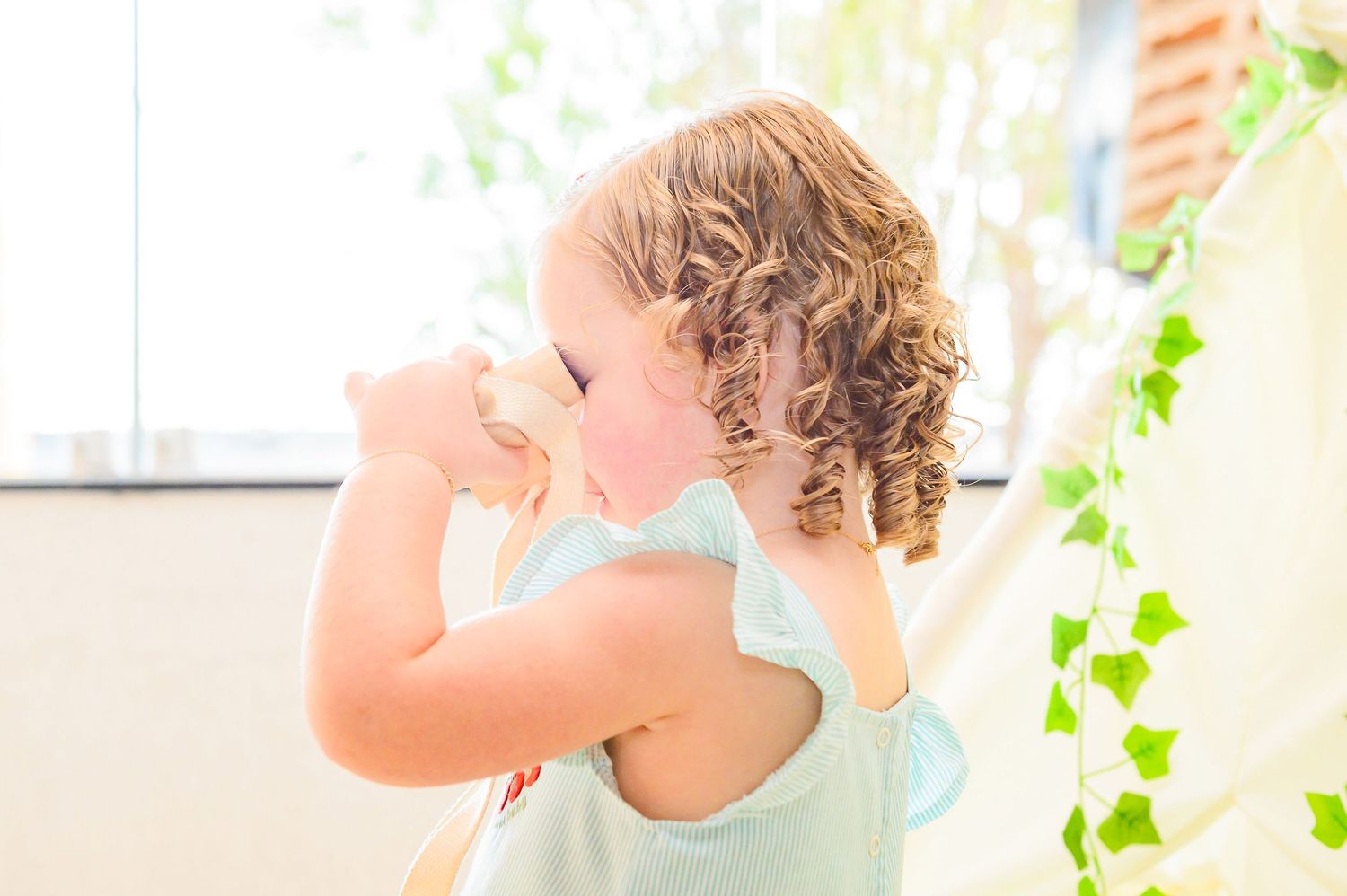 Small curly-haired toddler in light blue tank top stands near window with green ivy vines in soft natural light.