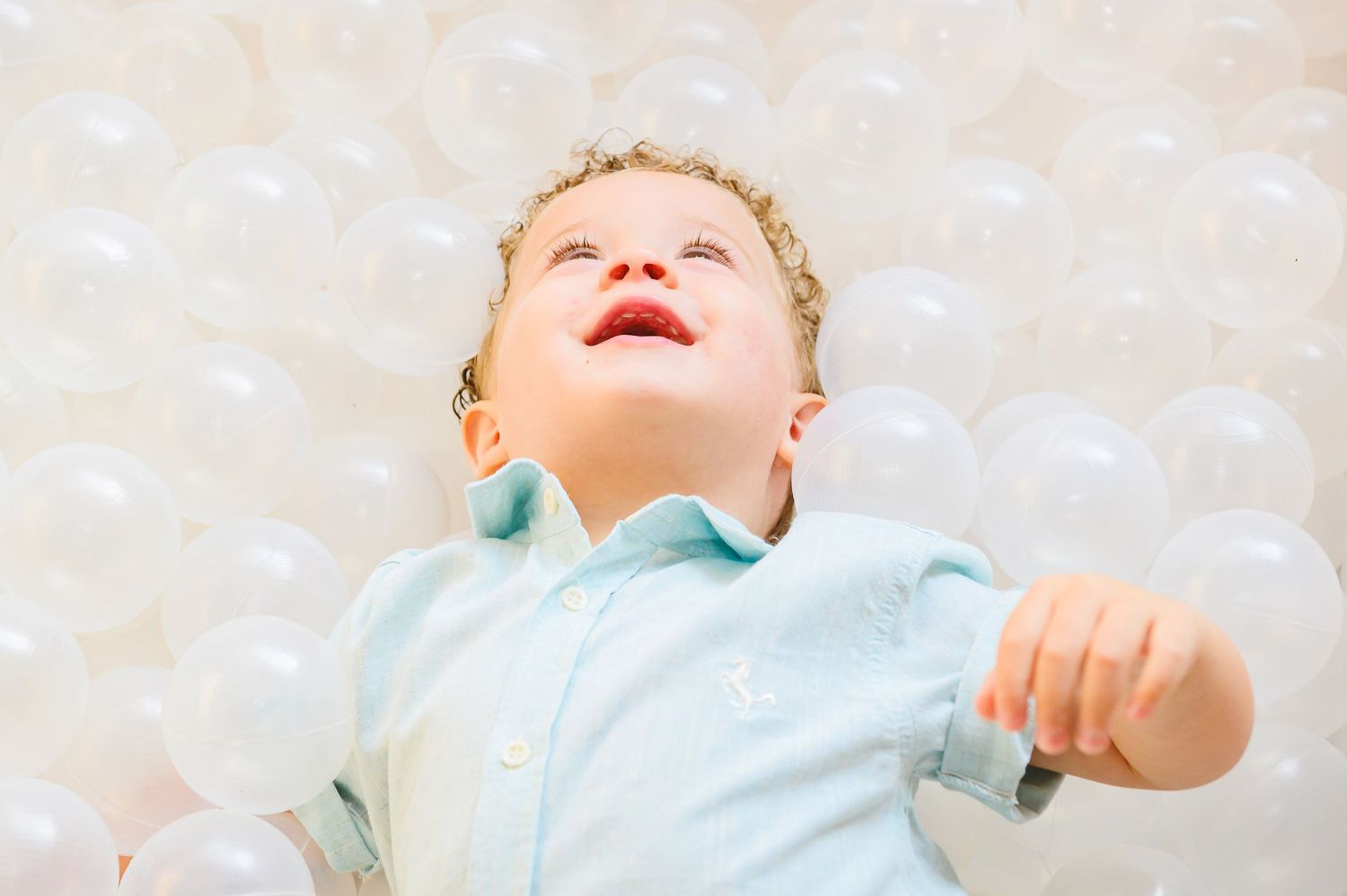 Young child in light blue shirt lies back looking up with joyful expression against soft light background.
