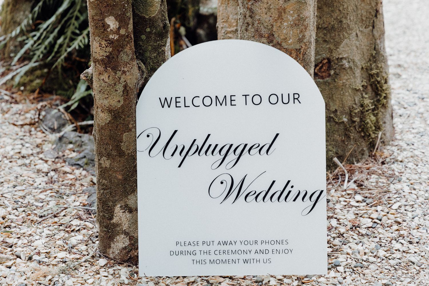 An elegant white arch-shaped wedding sign 'Welcome to Our Unplugged Wedding' captured by Apertured Media Auckland Wedding Photographer.