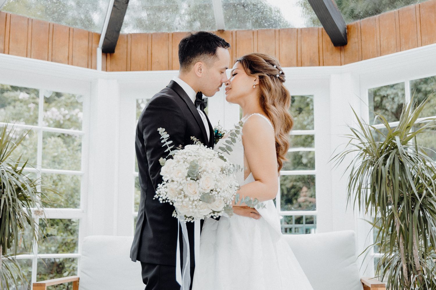 A couple shares a romantic moment in a sunlit conservatory with white flowers and tropical plants.