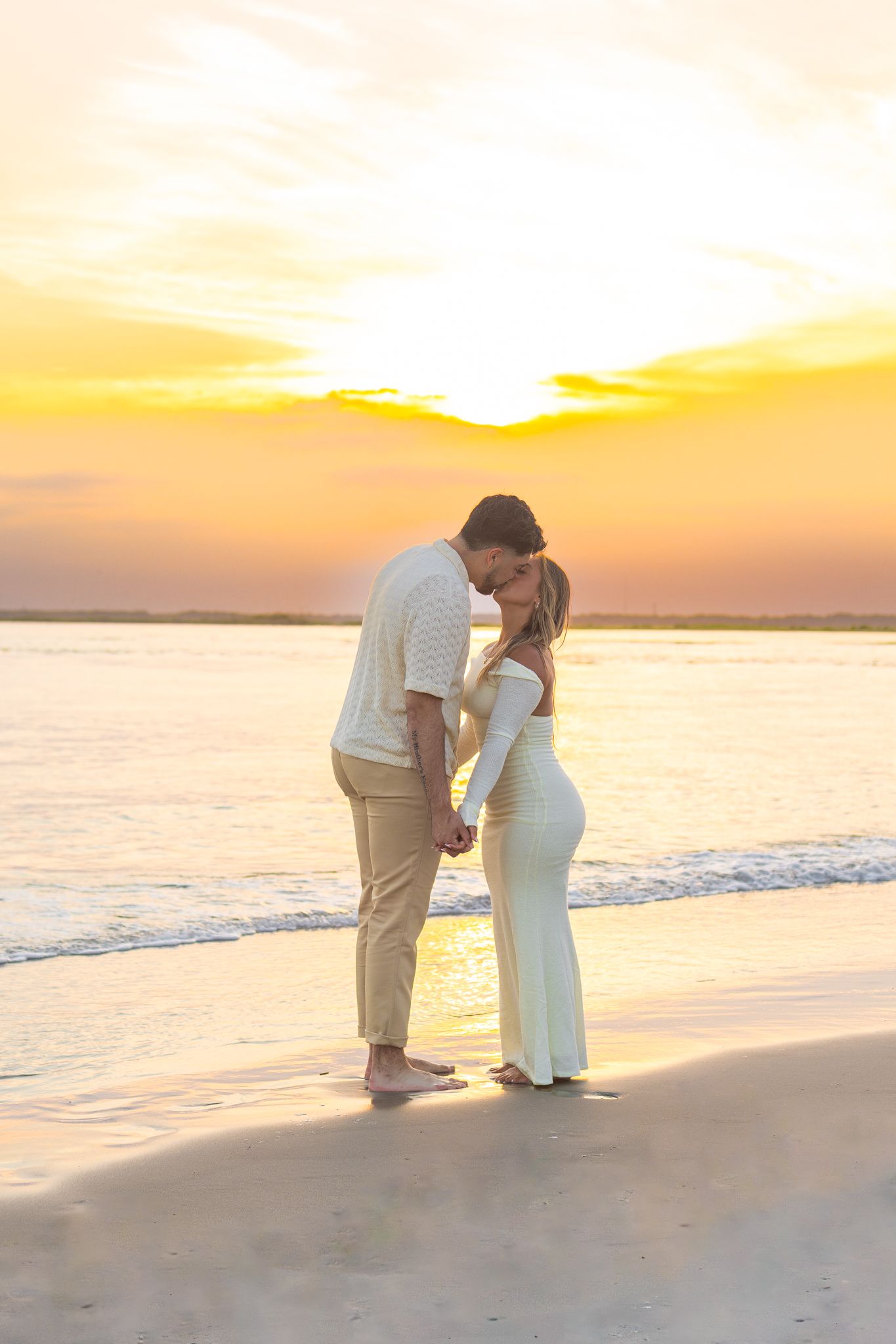 Romantic couple embraces on beach at sunset with golden sky and ocean waves in background.