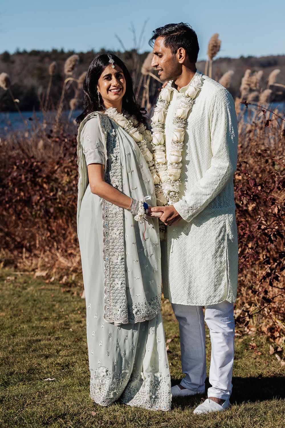 A couple stands outdoors wearing traditional white Indian wedding attire with floral garlands by a lake.