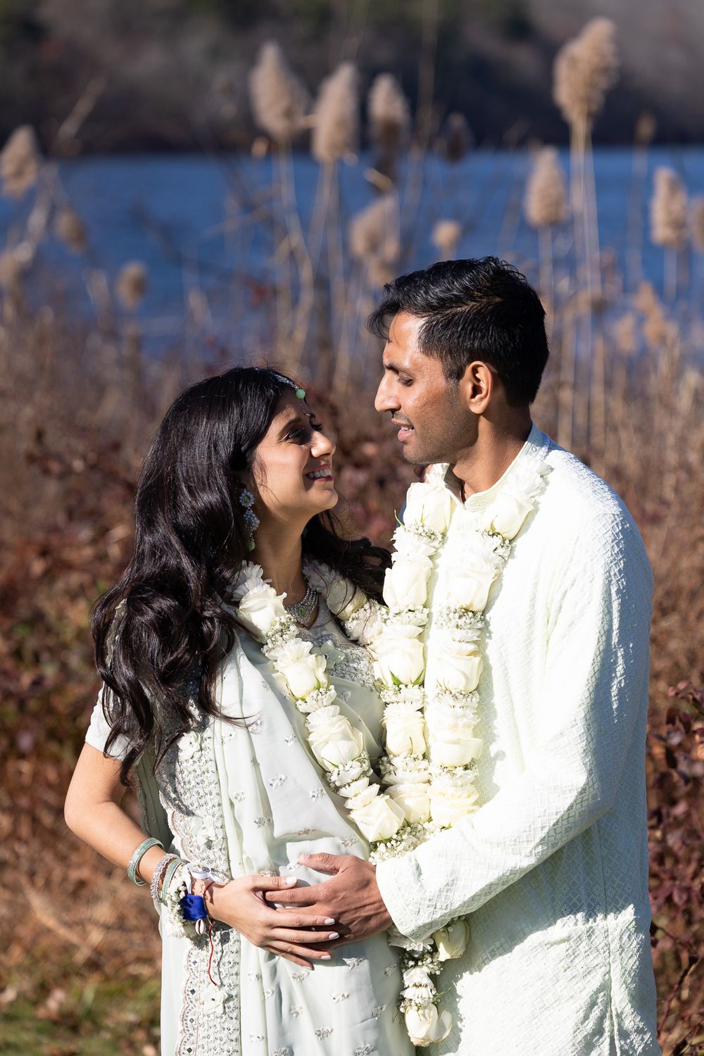 A couple in white traditional Indian attire share an intimate moment outdoors near cattails and water.