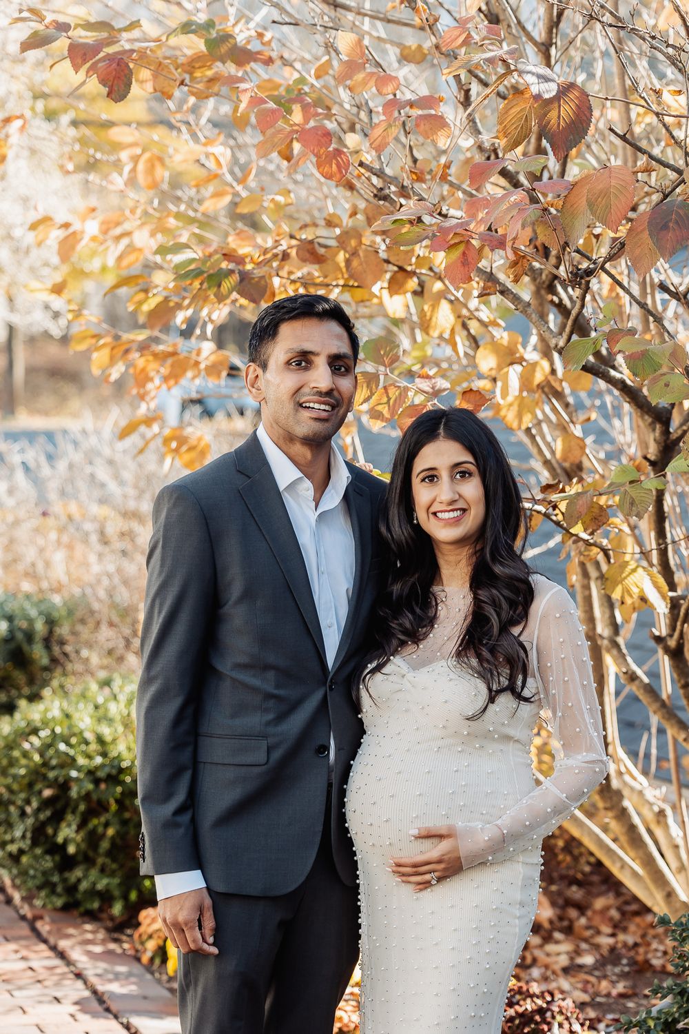 A couple poses together outdoors with autumn foliage, showing off her white fitted maternity dress.