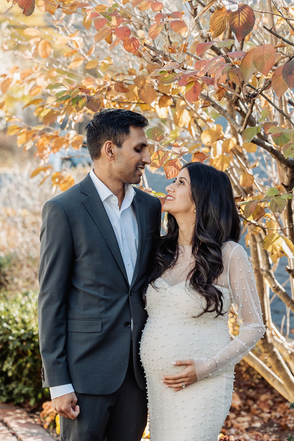 A couple poses intimately under fall foliage, she wearing a fitted white dress highlighting her pregnancy.