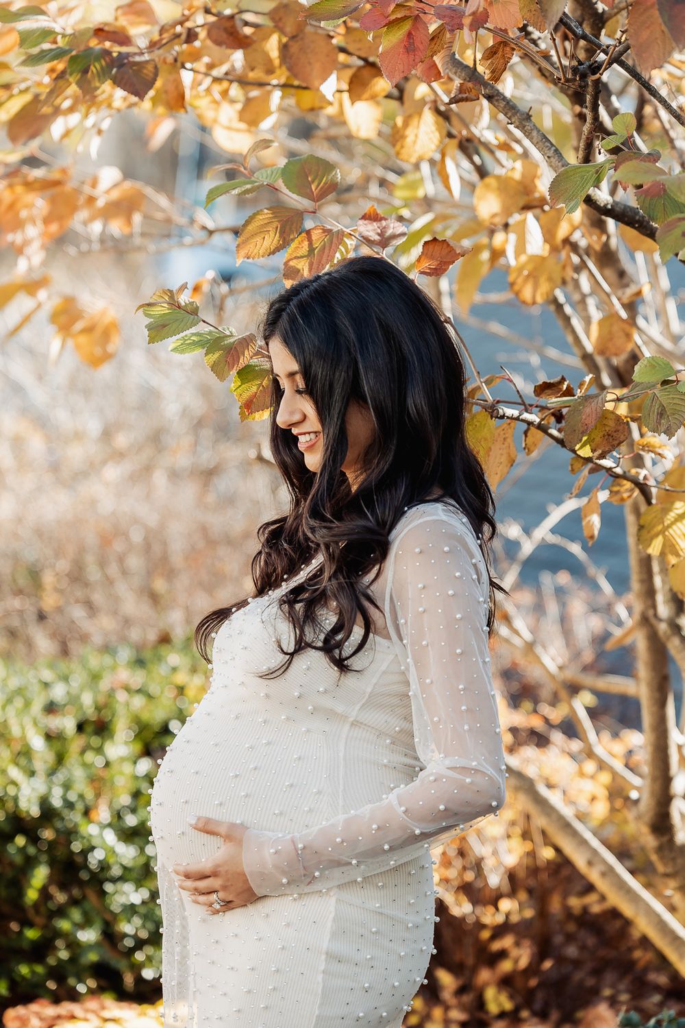 Woman in white long-sleeve maternity dress poses in profile under fall foliage.