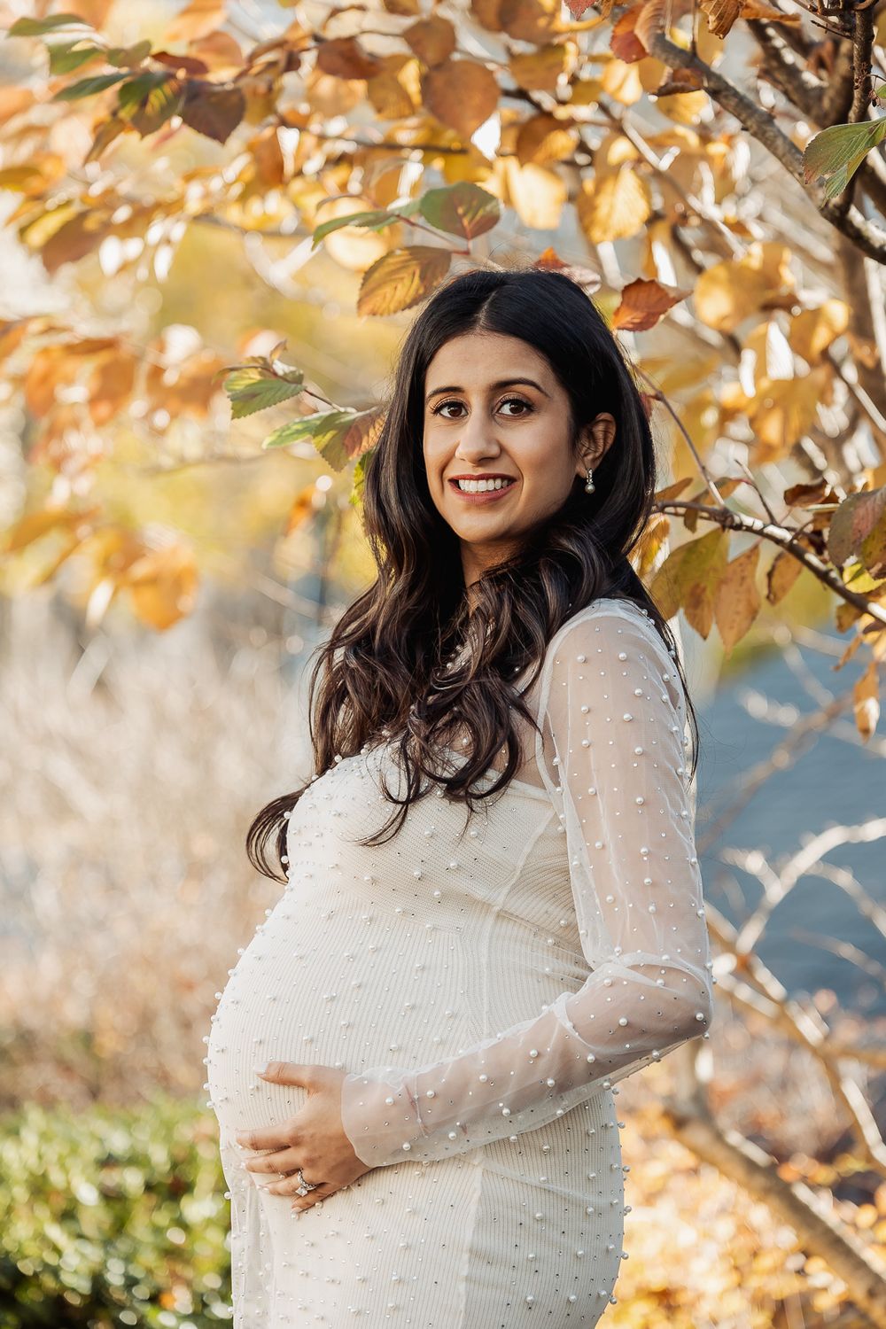 A woman in a white pearl-studded dress poses outdoors under golden autumn leaves.
