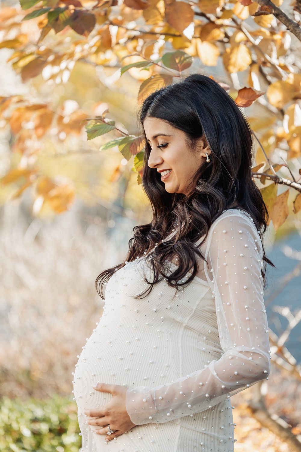 Expecting mother in white dress smiles while cradling her baby bump under golden autumn leaves.
