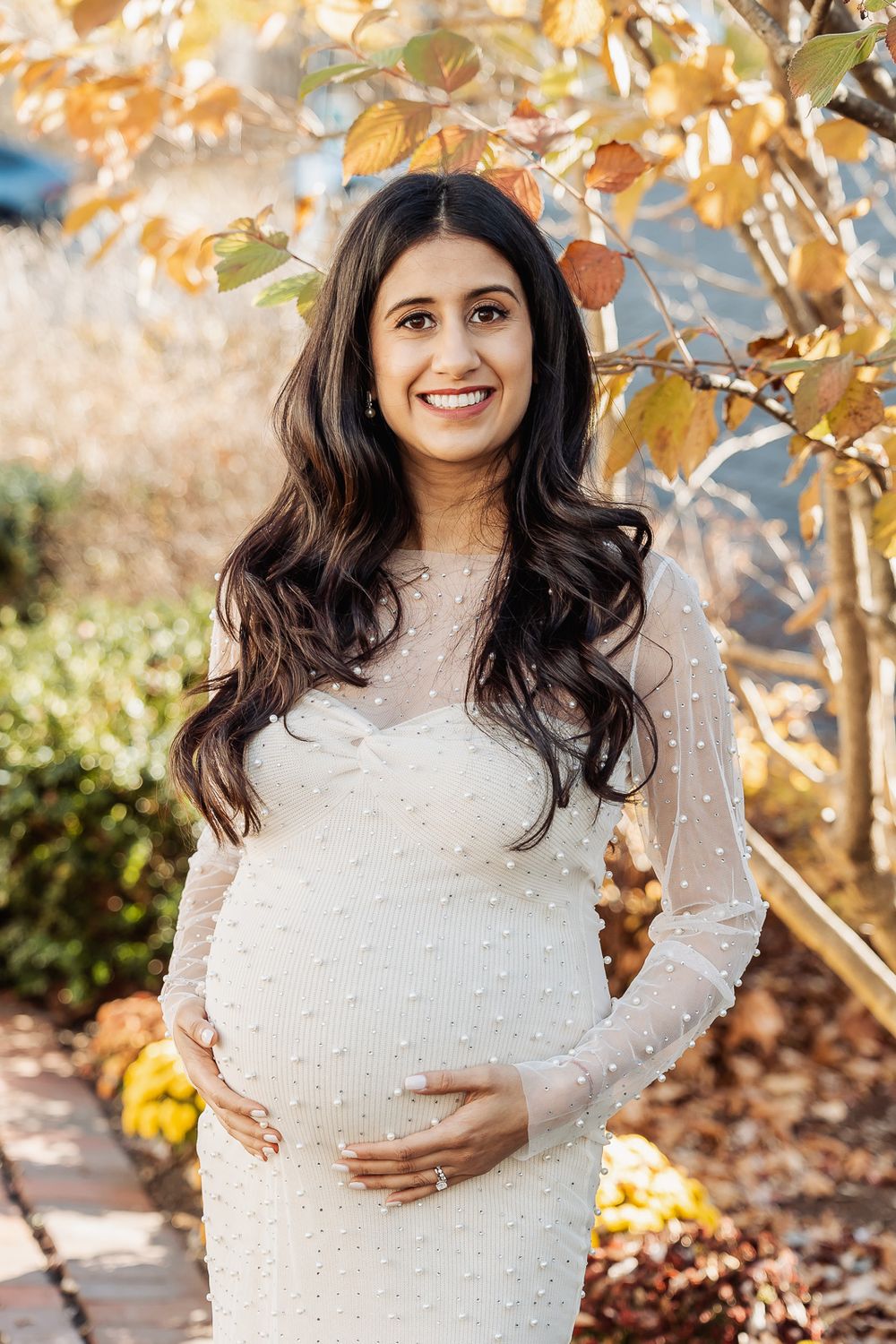 Woman in white pearl-embellished dress poses outdoors for maternity photos with fall foliage.