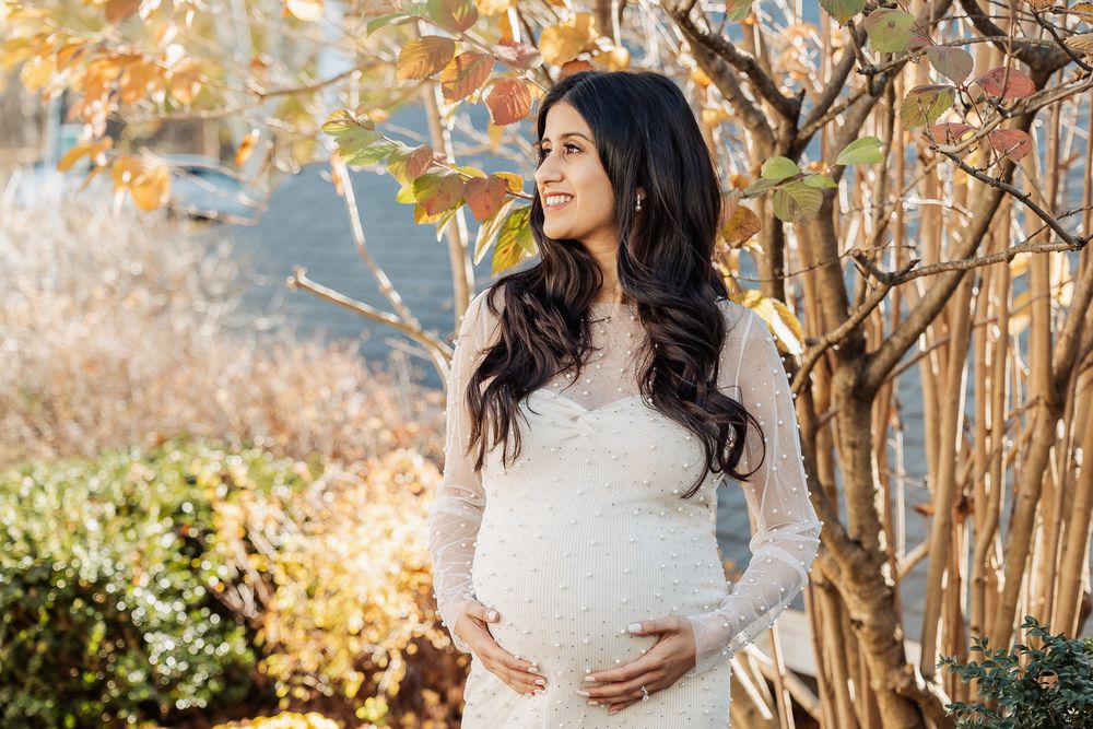 Expectant mother in long sleeve white dress poses by autumn foliage and garden.