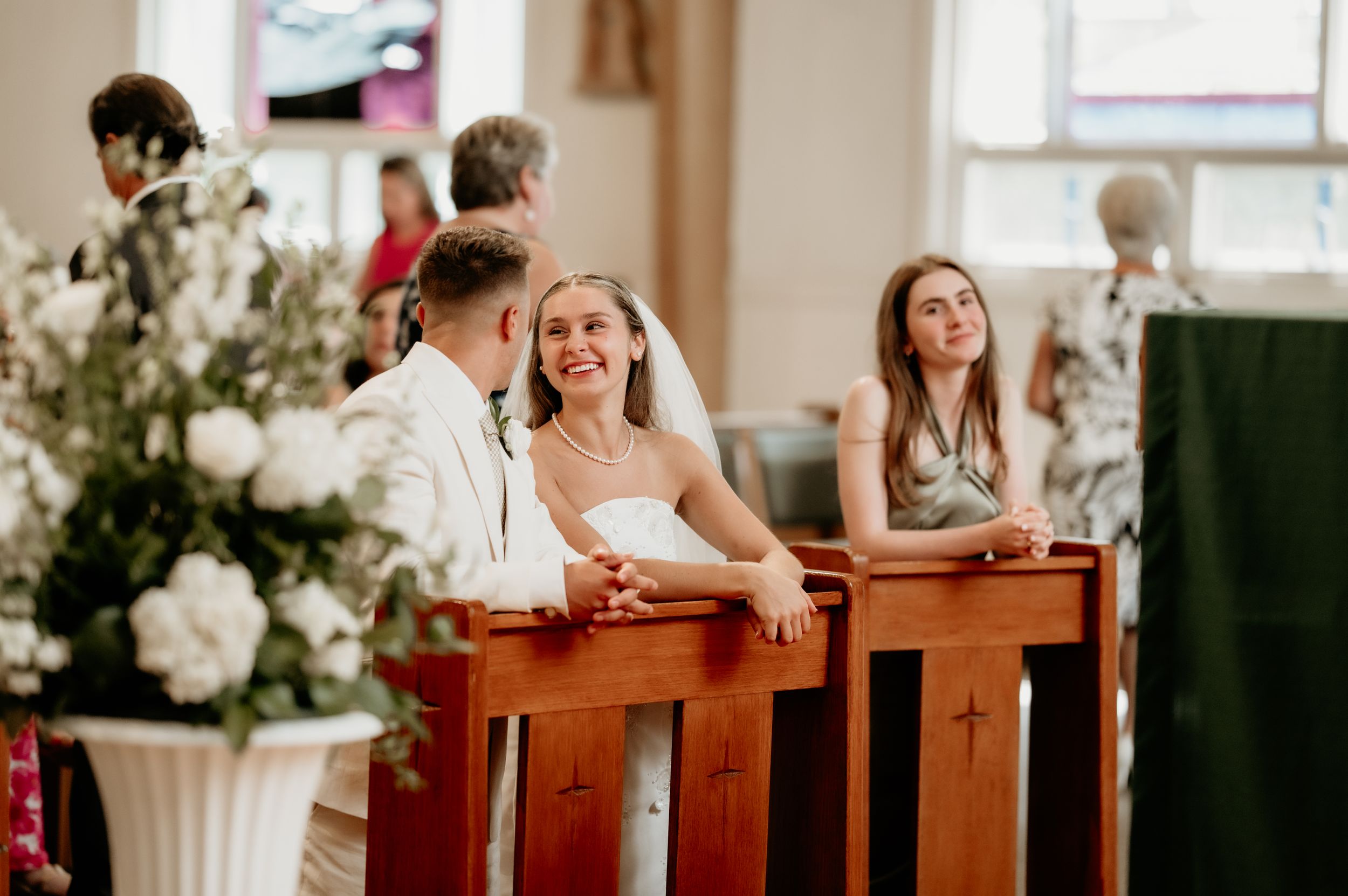 Wedding guests share laughter while seated in wooden church pews decorated with white flowers.
