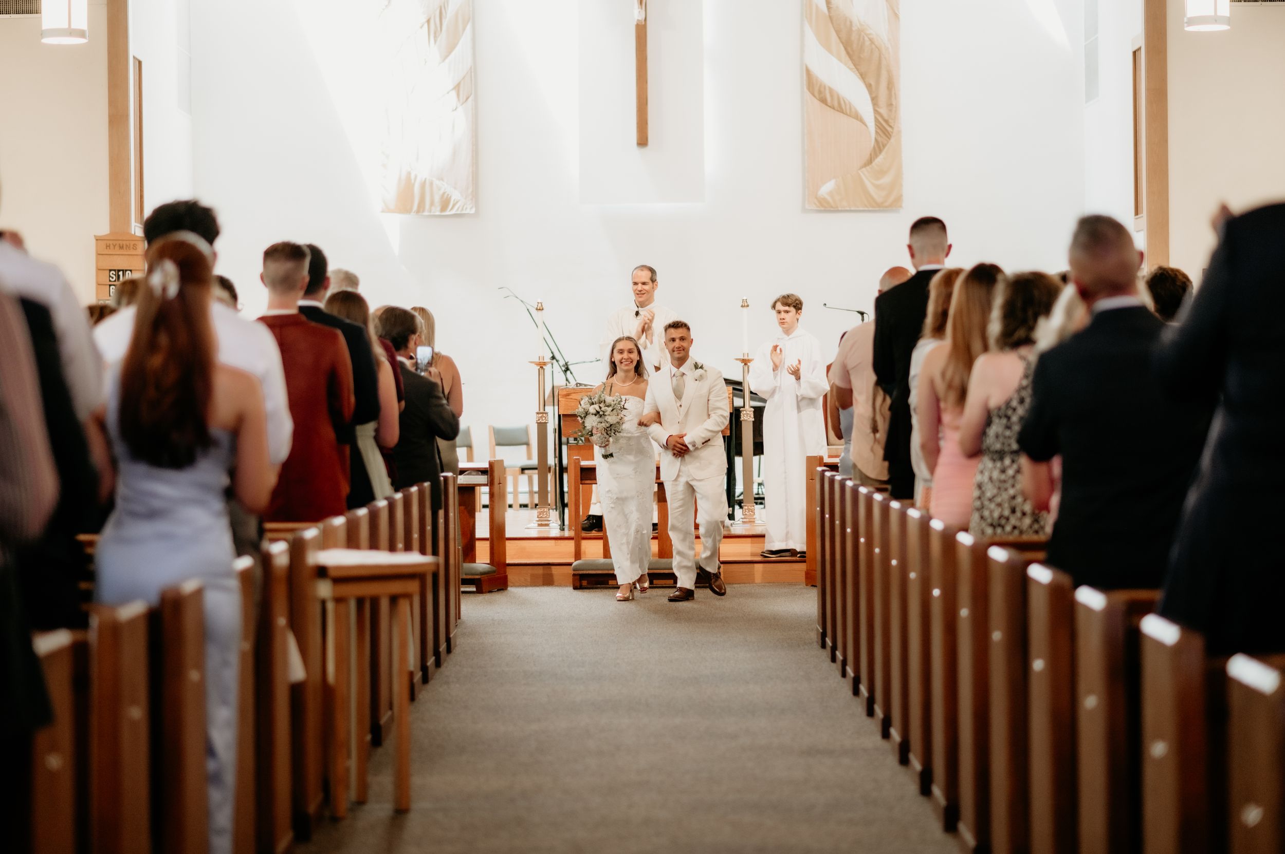 View down church aisle during wedding ceremony with guests seated in traditional wooden pews.