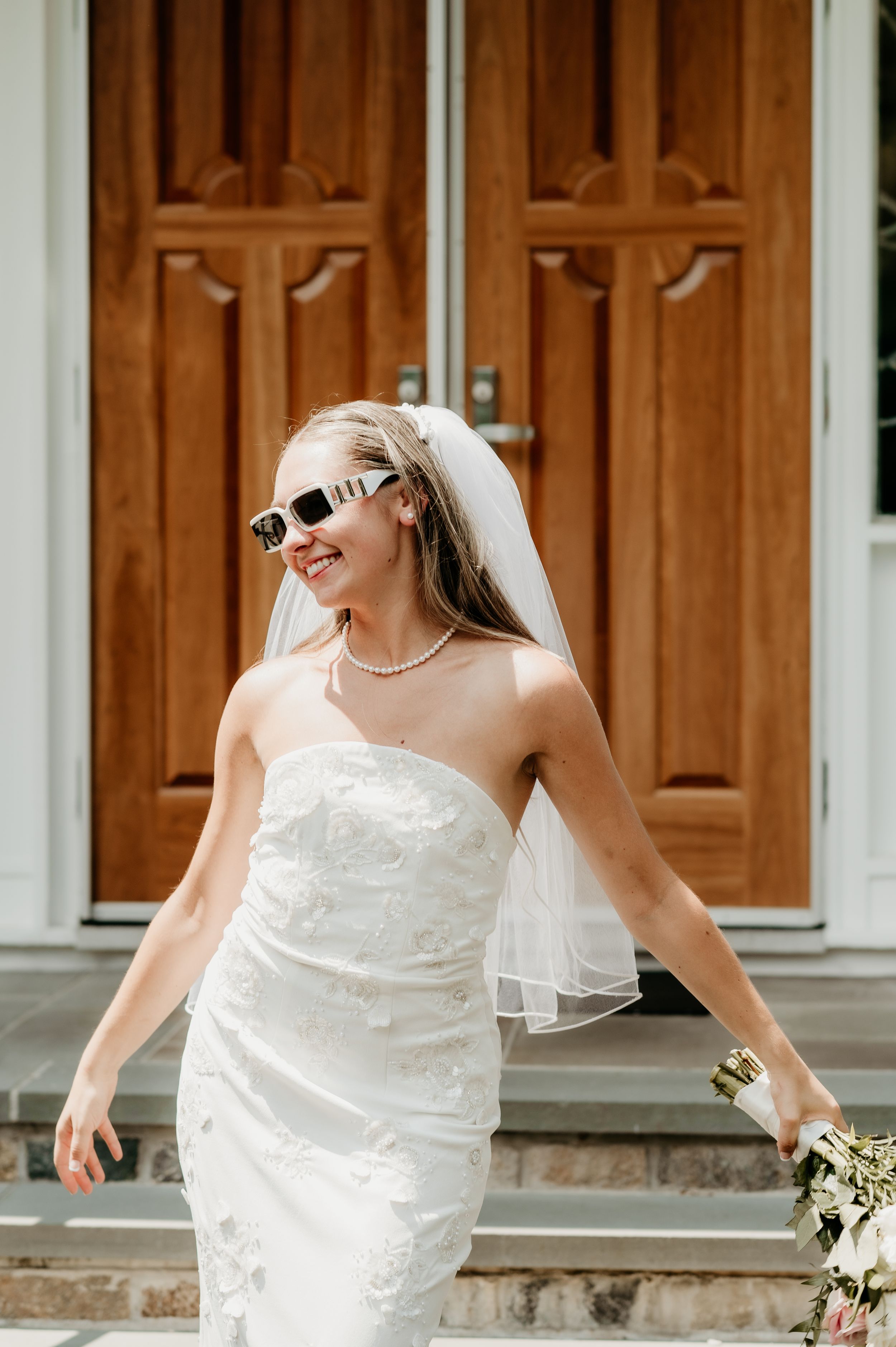 Bride wearing sunglasses and strapless wedding dress poses playfully in front of wooden church doors.