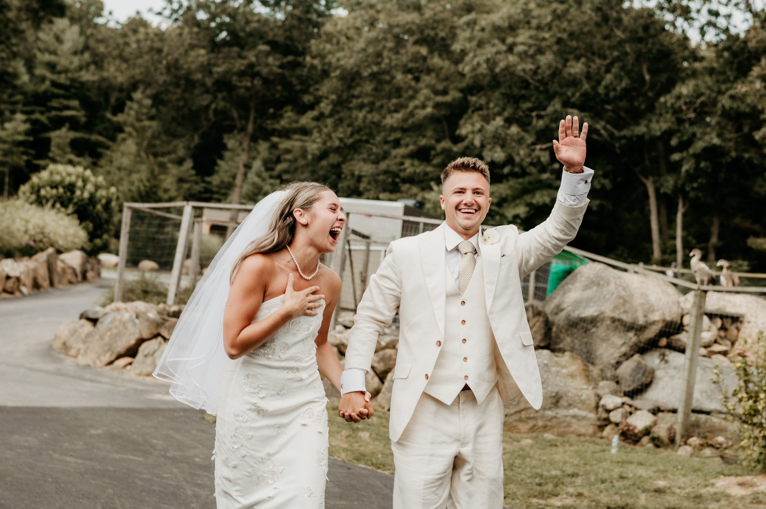 Newlyweds in white attire celebrate joyfully while walking hand-in-hand outdoors. Rhode Island.