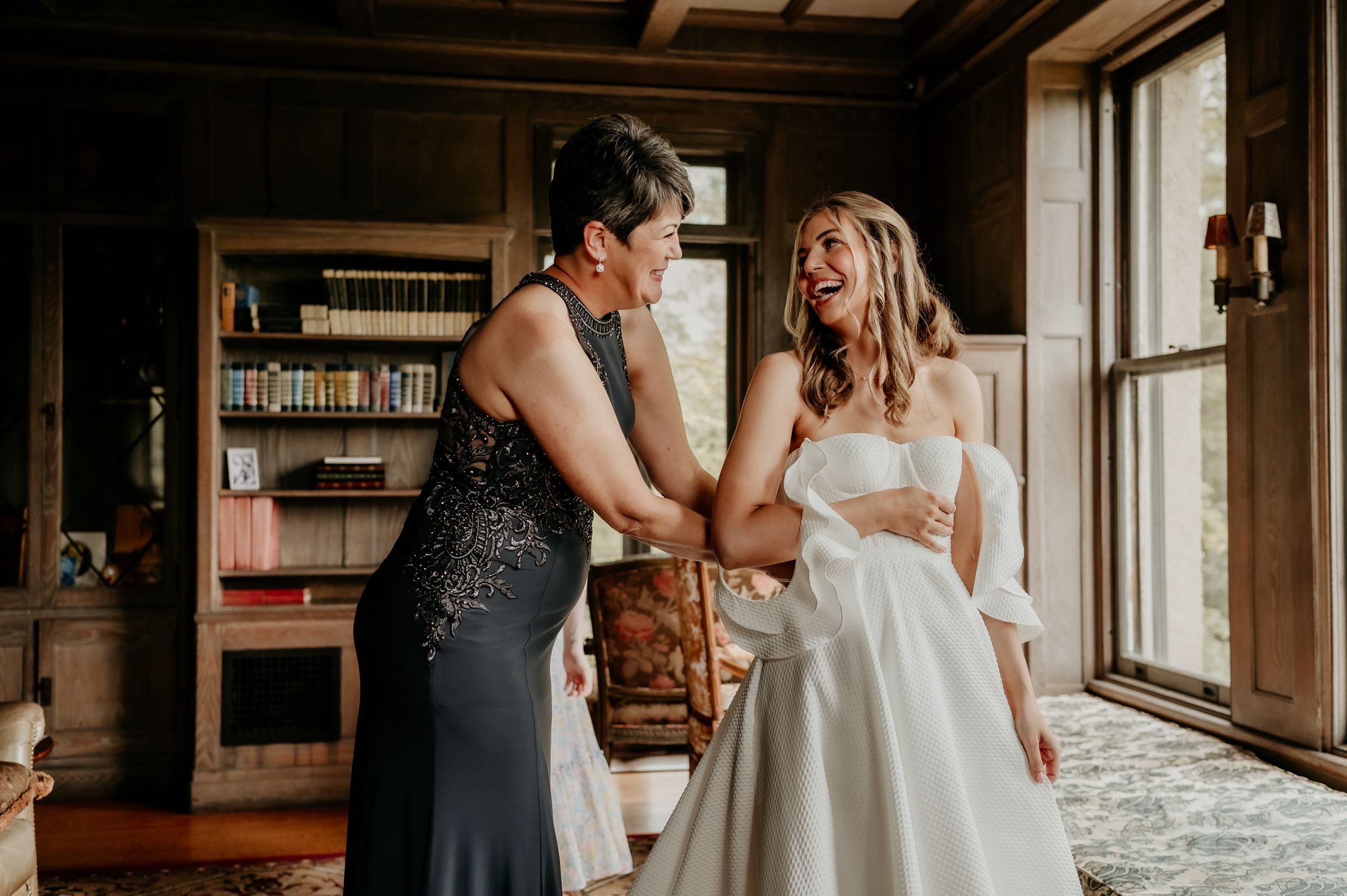 Two elegantly dressed women share an intimate moment in a library setting during a wedding celebration.