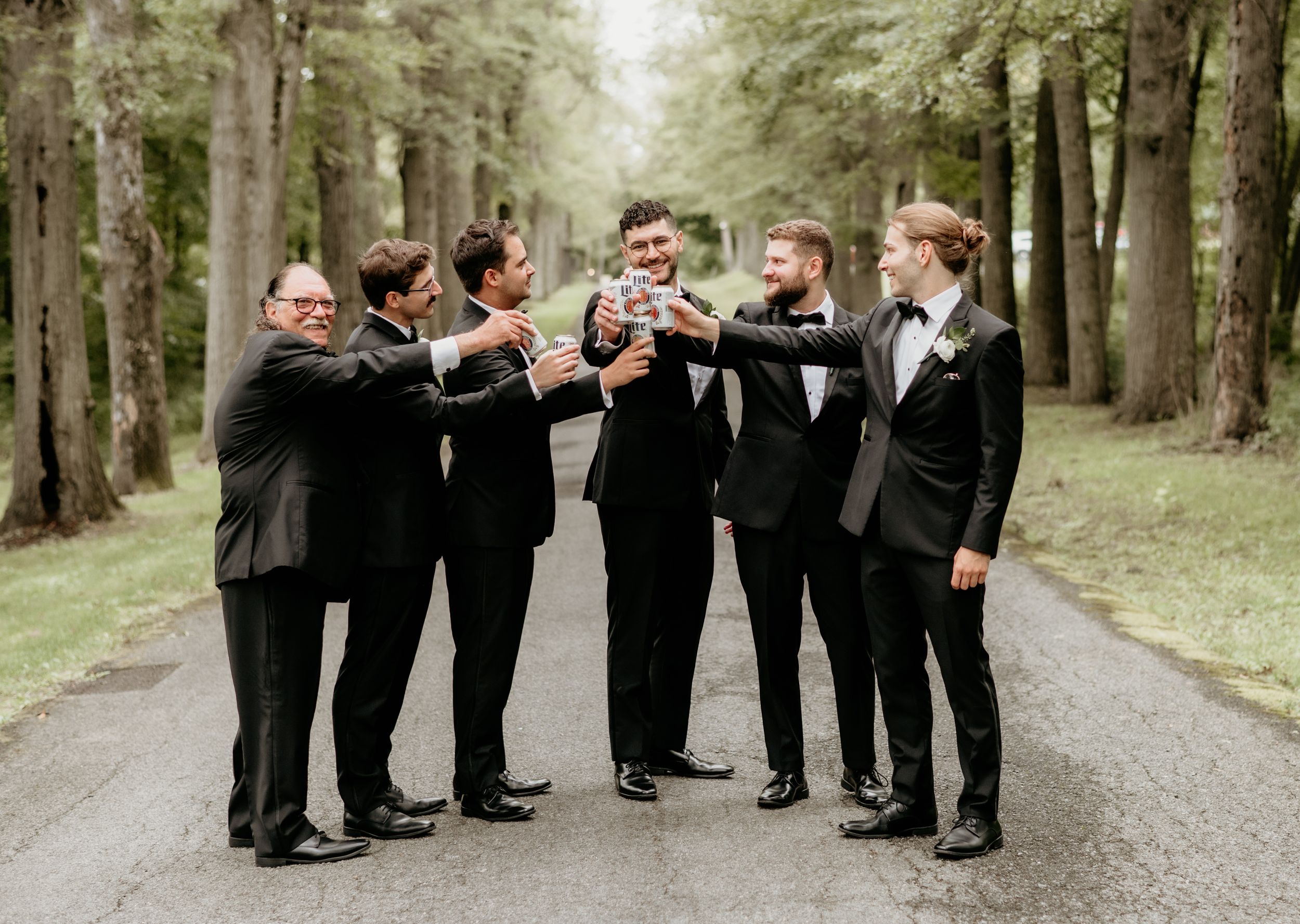 Groomsmen in black tuxedos toast and celebrate together on a tree-lined path.