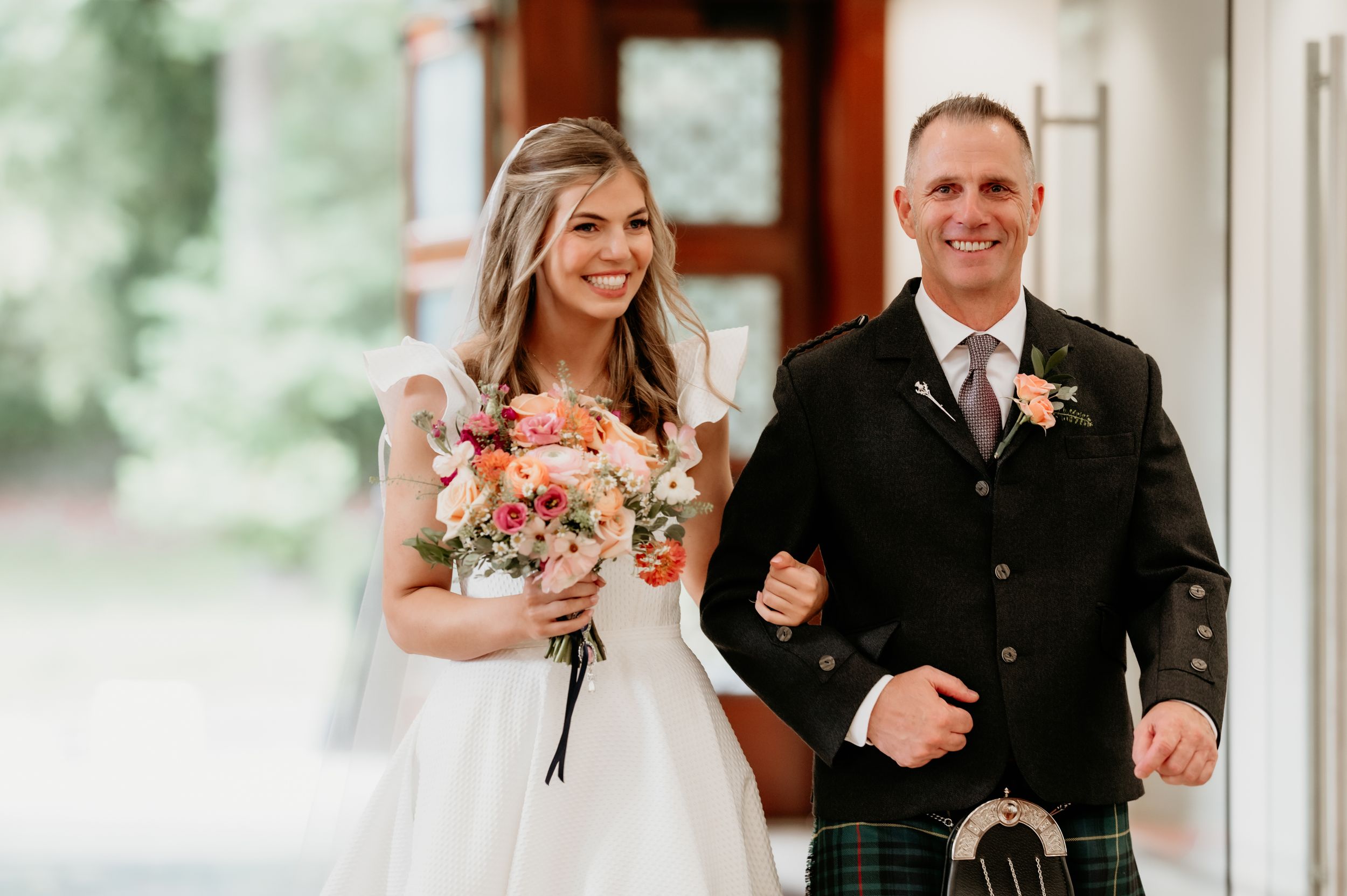 A bride in white dress holds pink bouquet while walking with escort in traditional Scottish kilt.