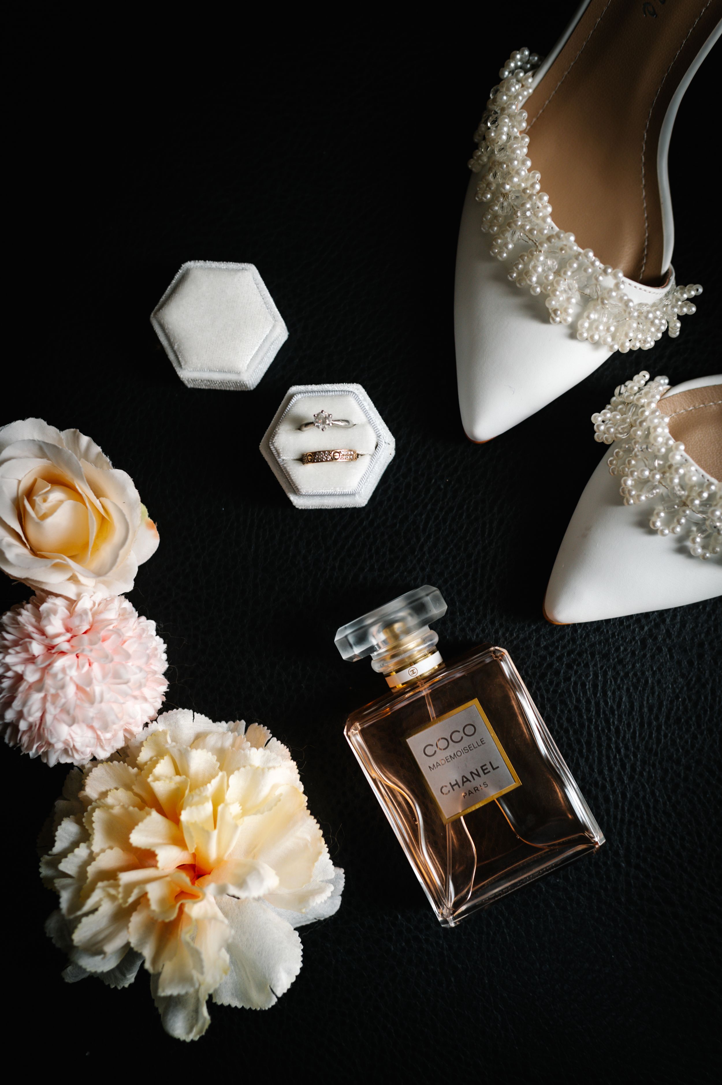 Wedding accessories flatlay with perfume, white shoes, rings and peach flowers on black surface.