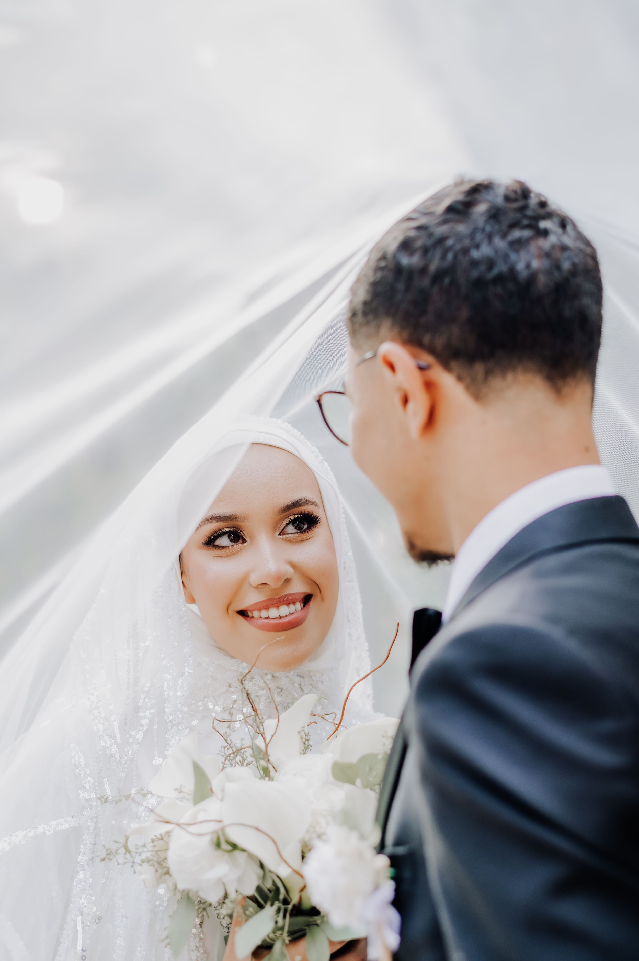Tender moment between newlyweds under flowing white veil with bridal bouquet.