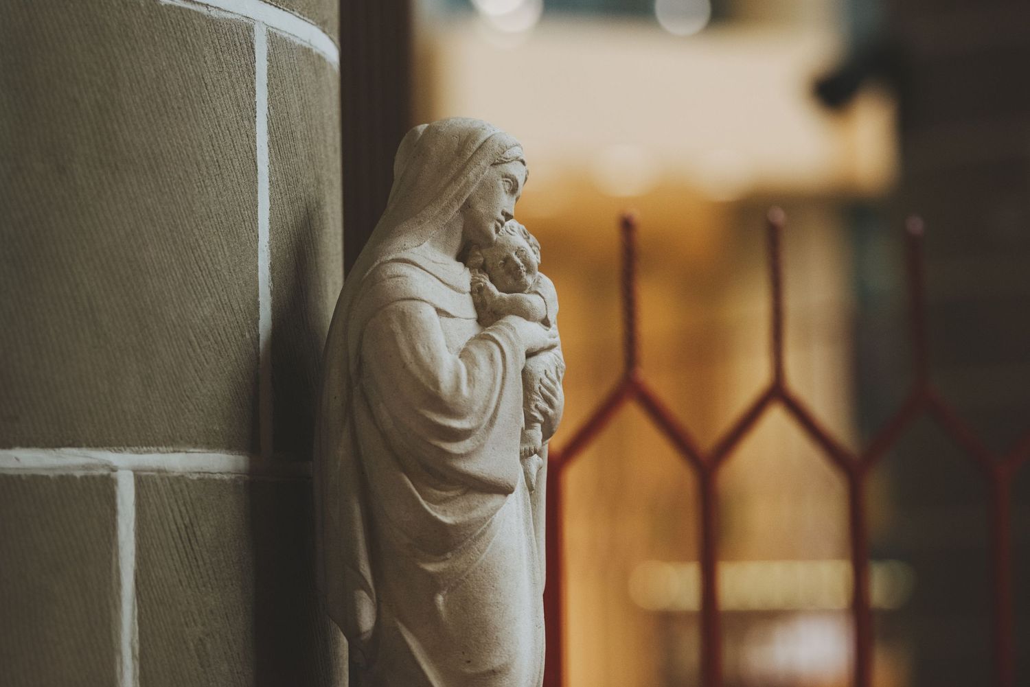 A figure of the virgin Mary and child stands against a stone wall in soft focus with gothic architectural details visible.