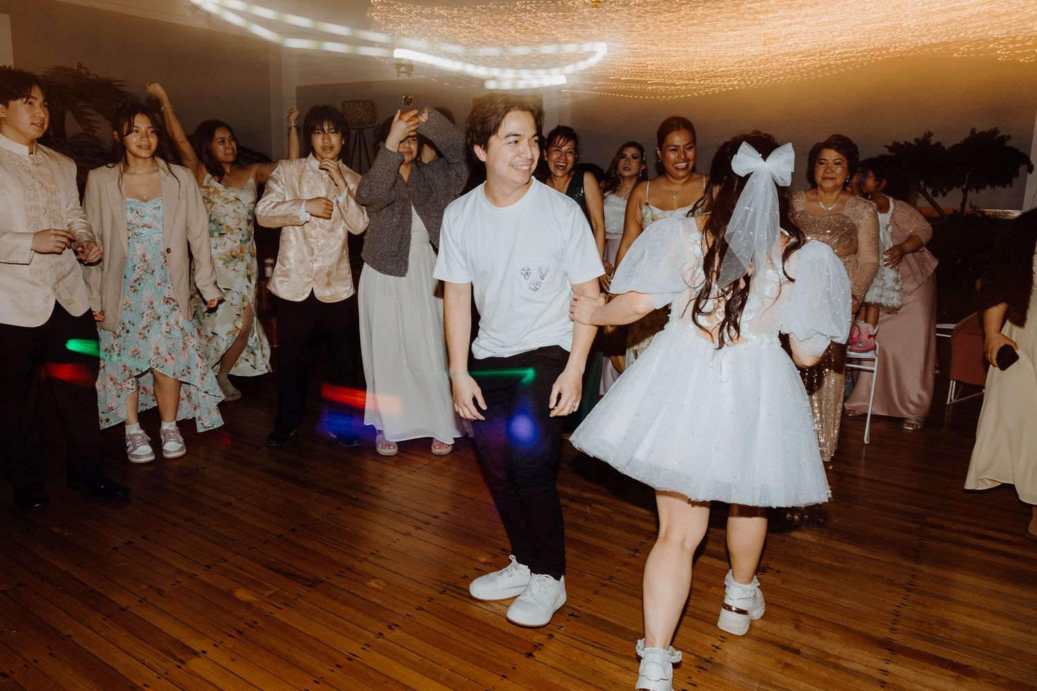 Wedding guests dancing in a reception hall with string lights and hardwood floors.