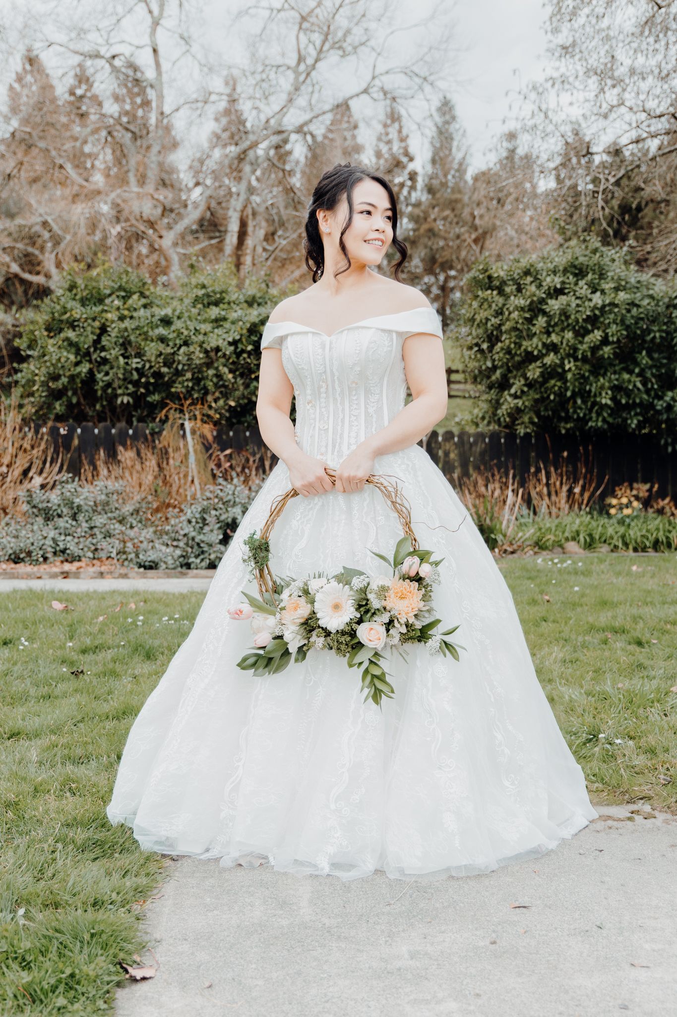 Bride in off-shoulder ballgown holding pastel floral bouquet in garden setting.