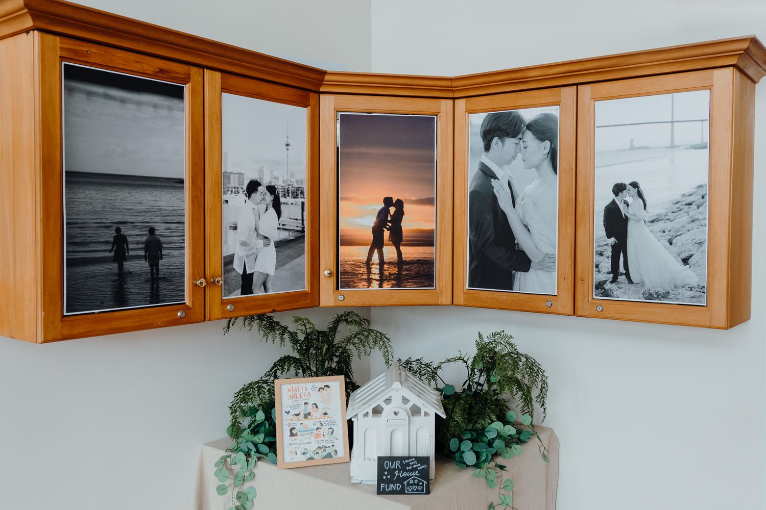 Black and white beach photos displayed in wooden corner cabinet with decorative greenery below.