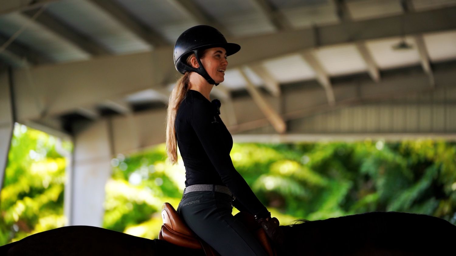 Equestrian rider in black attire practicing dressage on horseback in a covered arena.