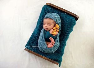 Sleeping baby wrapped in teal blanket on rustic brown backdrop holding small sunflowers.