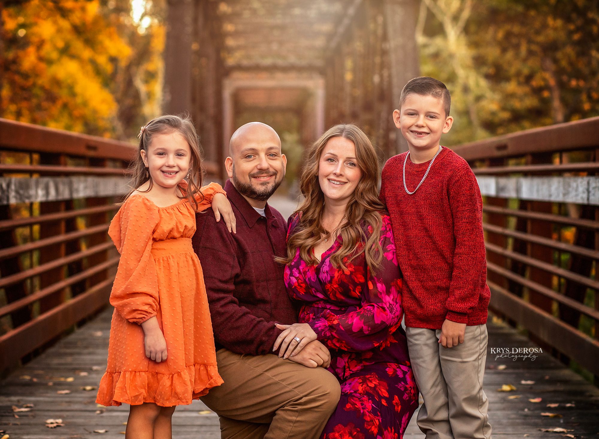 A family in coordinating autumn outfits poses together on a wooden bridge surrounded by fall foliage.