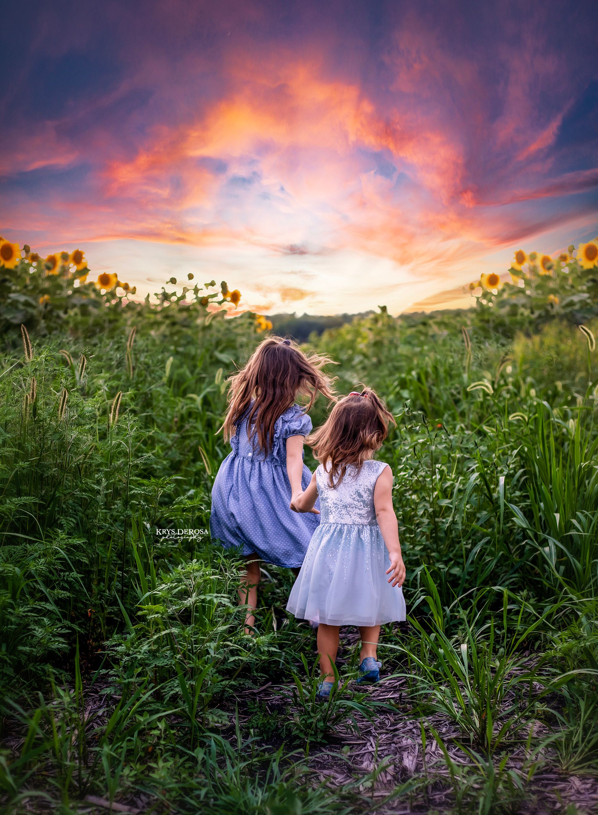 Two children in purple dresses walking through a sunflower field at sunset with vibrant orange and pink sky.