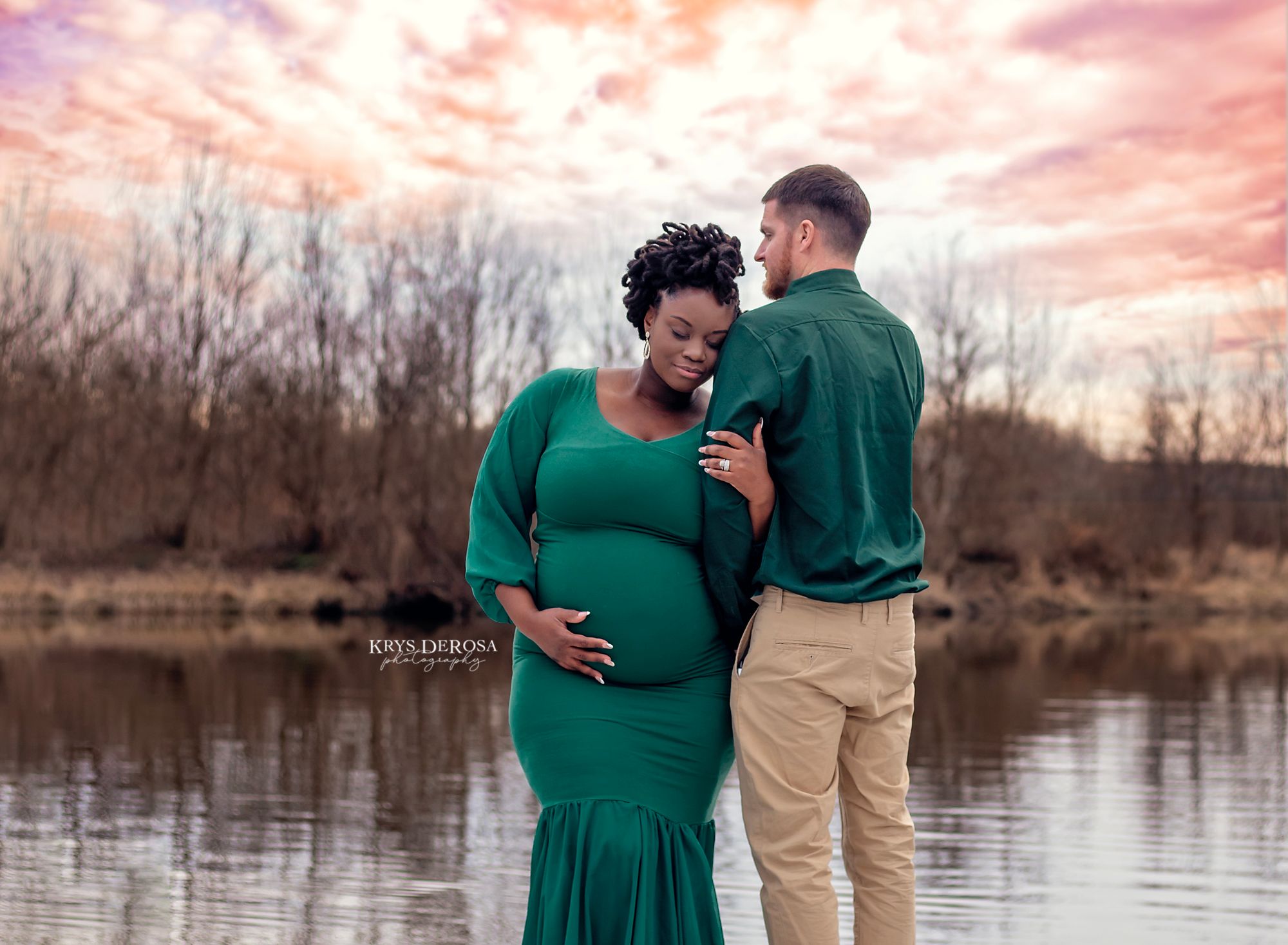 A couple in matching green outfits share an intimate moment by a lake at sunset with beautiful pink sky.