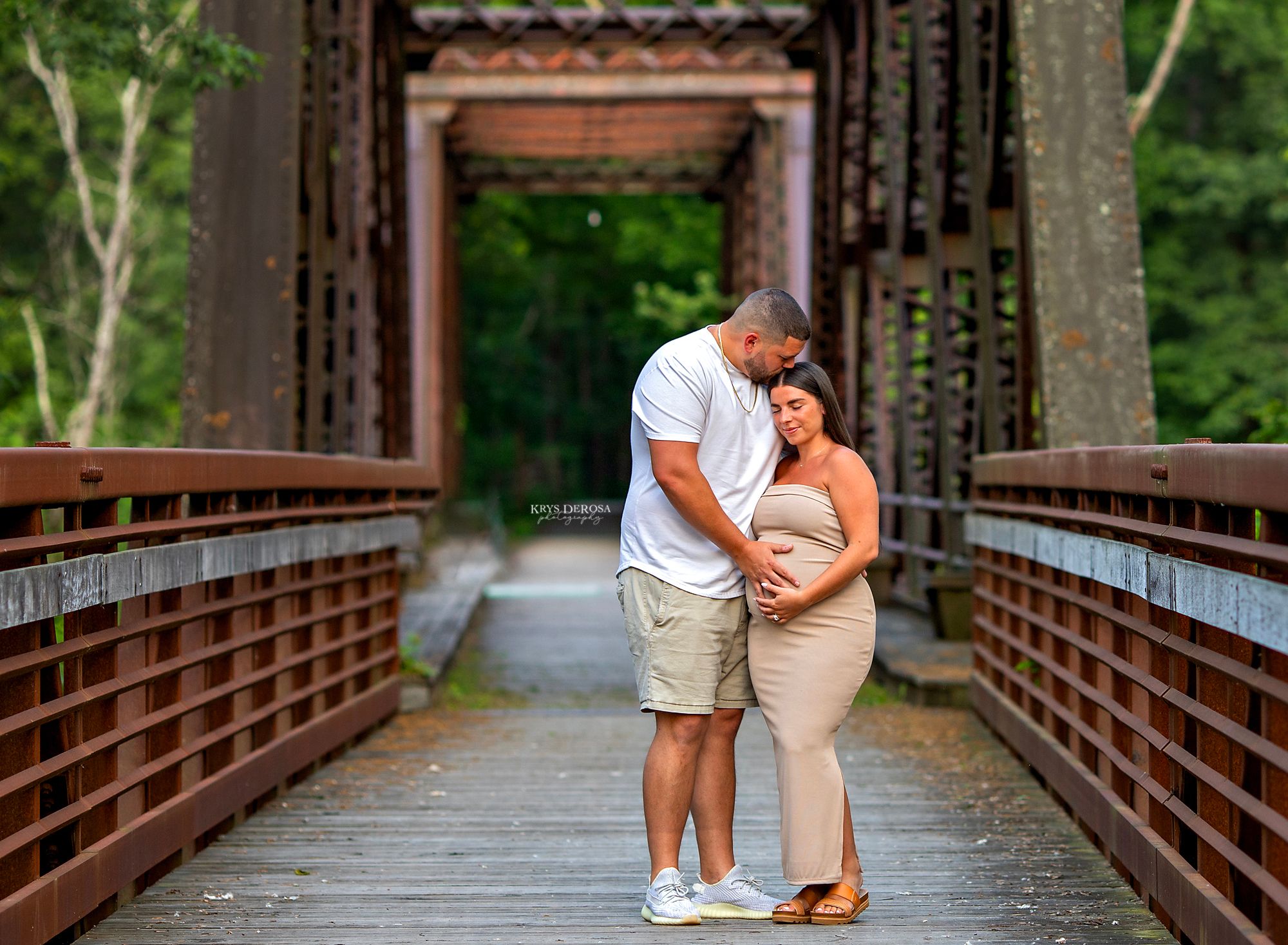A couple shares a romantic moment standing together on a rustic wooden and metal bridge surrounded by greenery.