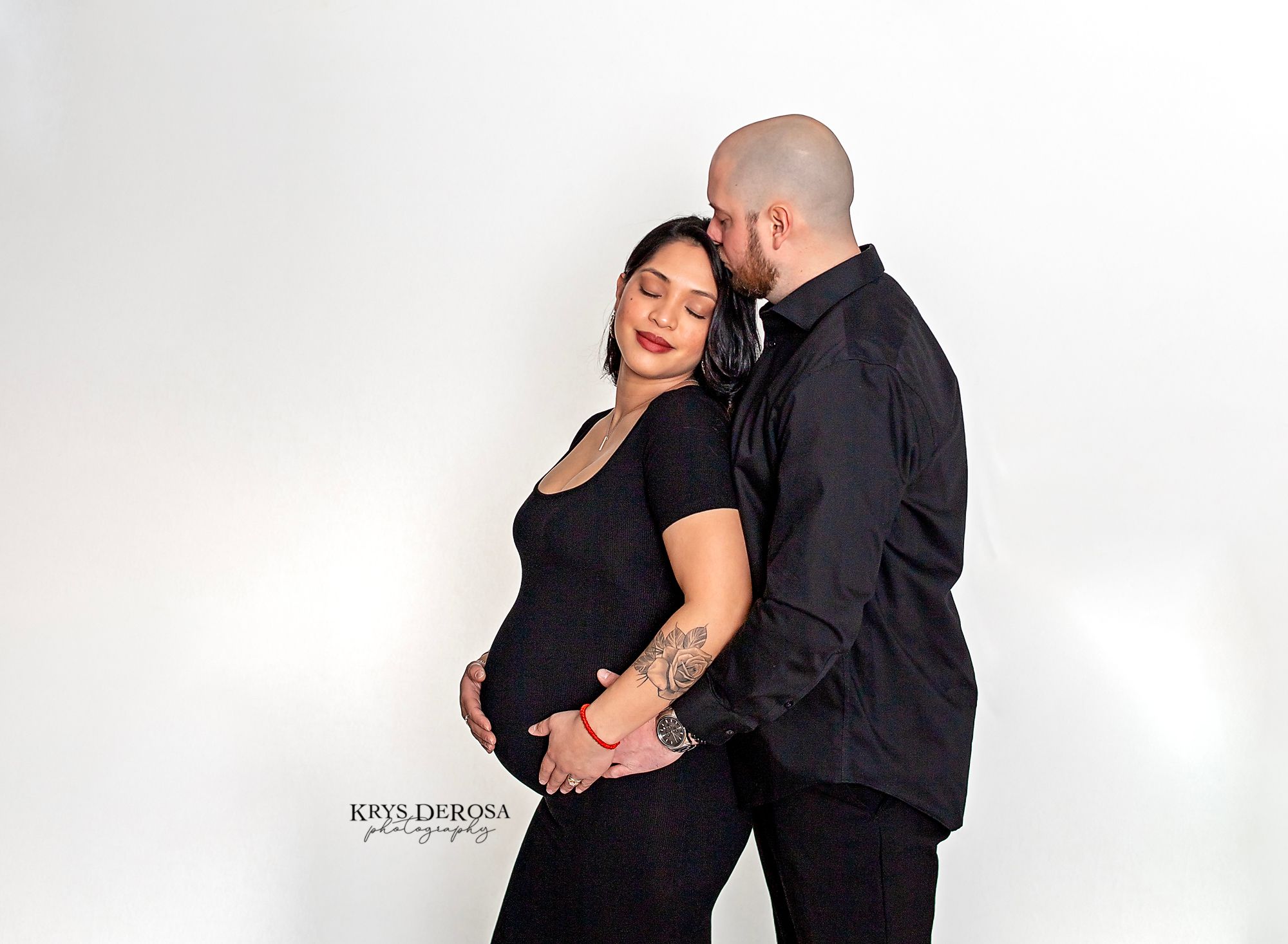 A couple dressed in black embraces against a white backdrop during an intimate studio portrait session.