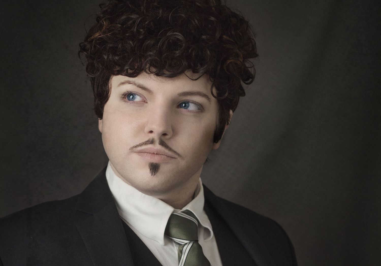 Professional portrait of a suited individual with dark curly hair and facial hair against a dark background.