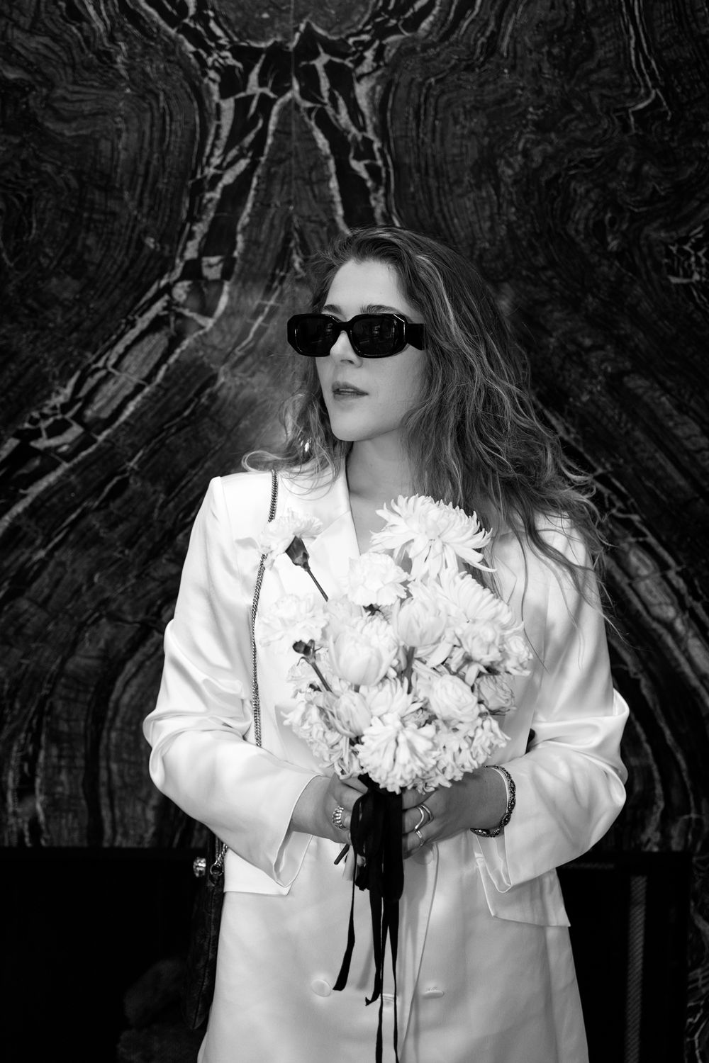 A figure in sunglasses and white coat holds a bouquet of white flowers against a dark textured background.