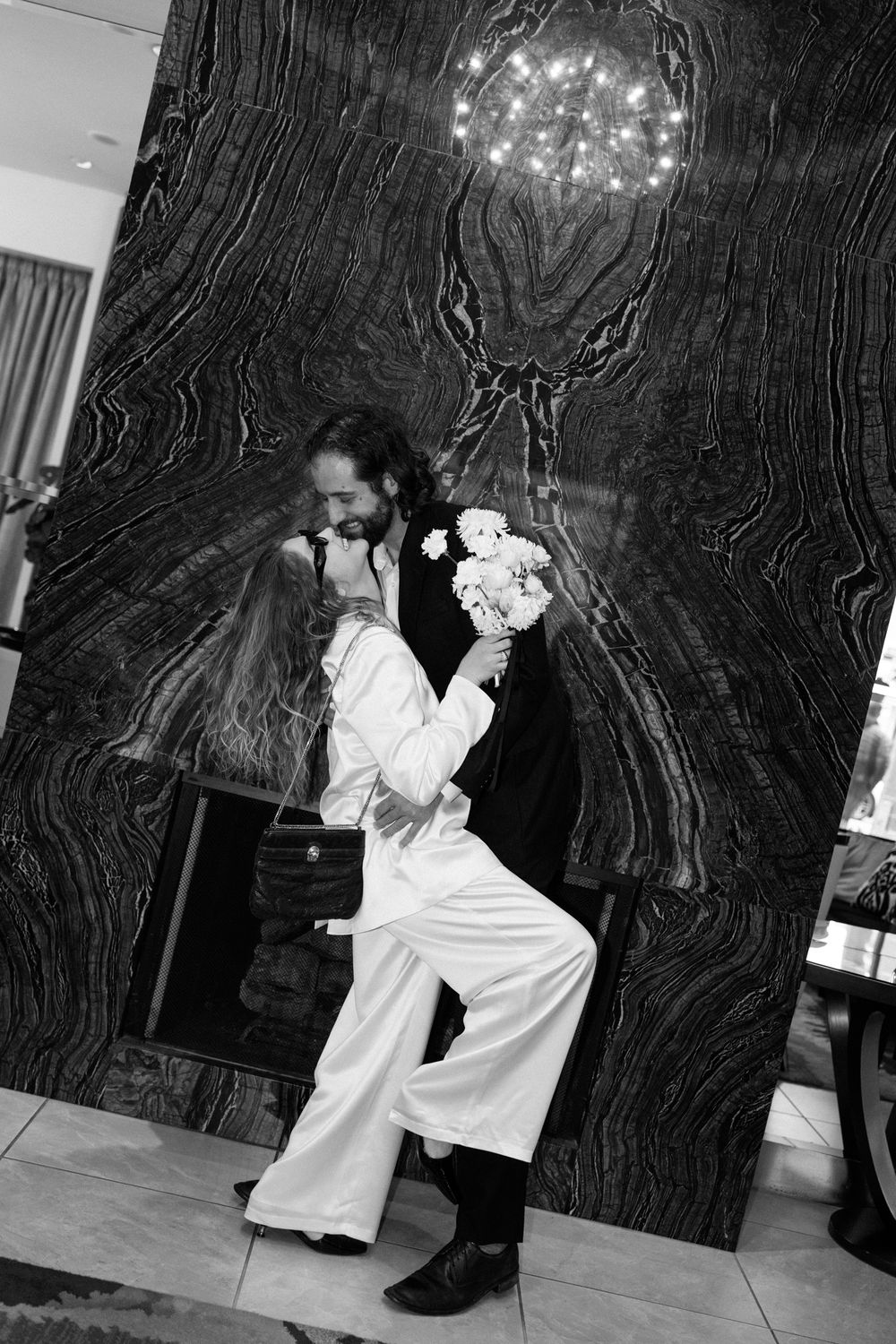 Black and white photo of a couple dancing intimately beneath a chandelier against a dark textured wall.