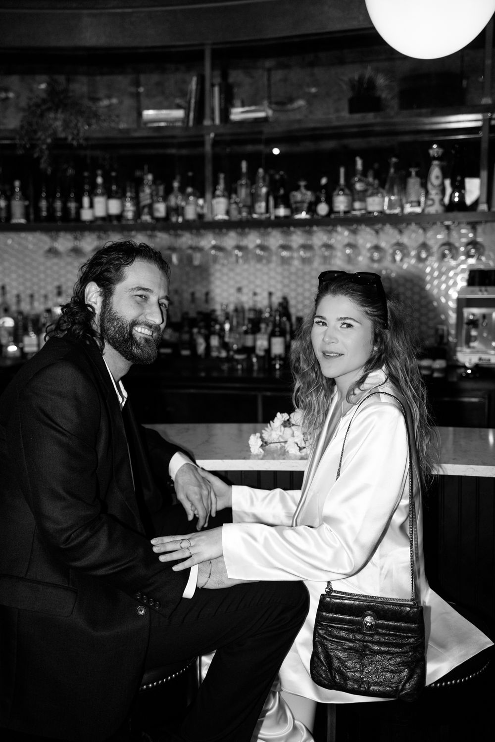 A couple enjoys drinks together at a bar with wine glasses and liquor bottles displayed on shelves in the background.