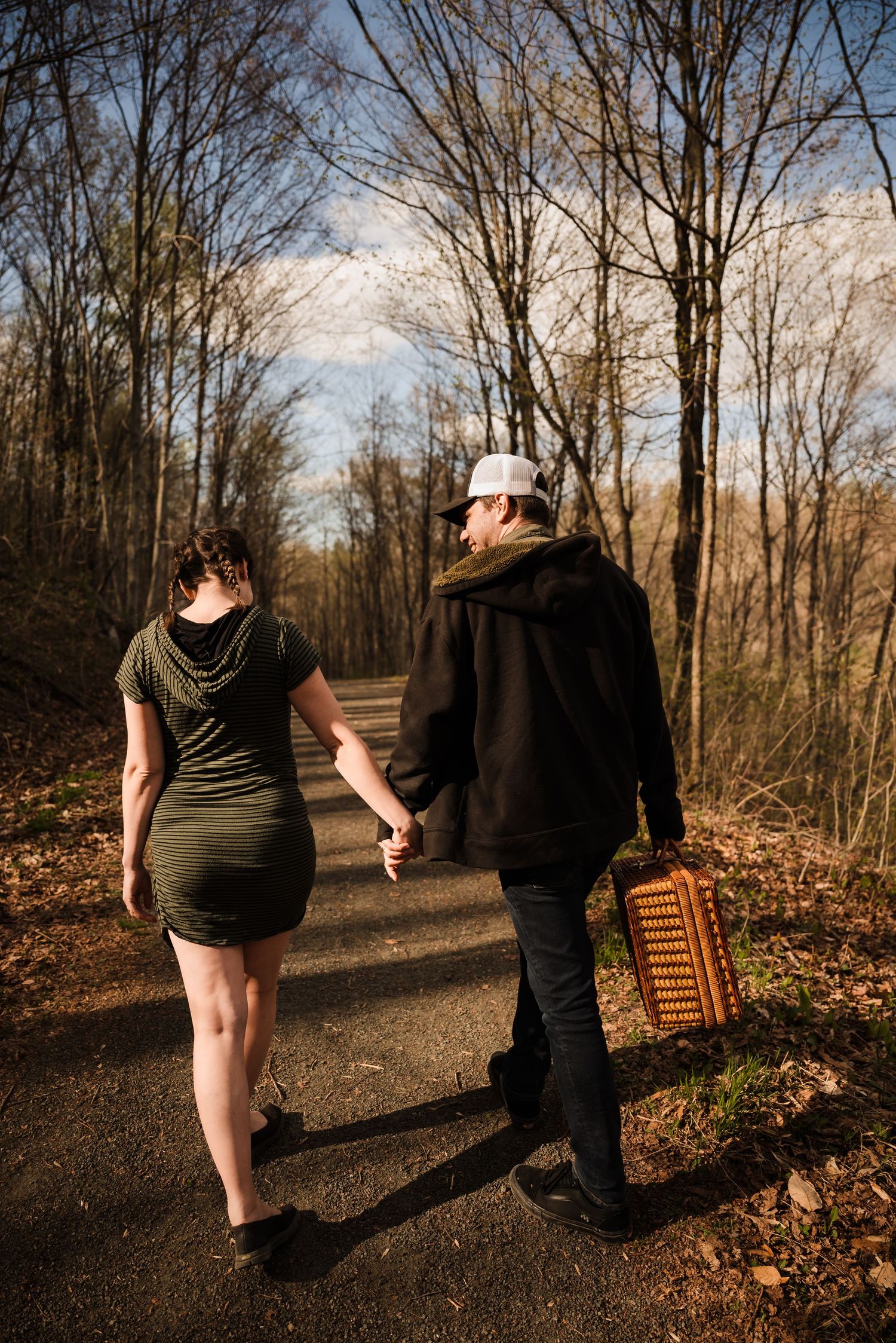 Couple walking hand in hand on forest trail with picnic basket during golden hour.