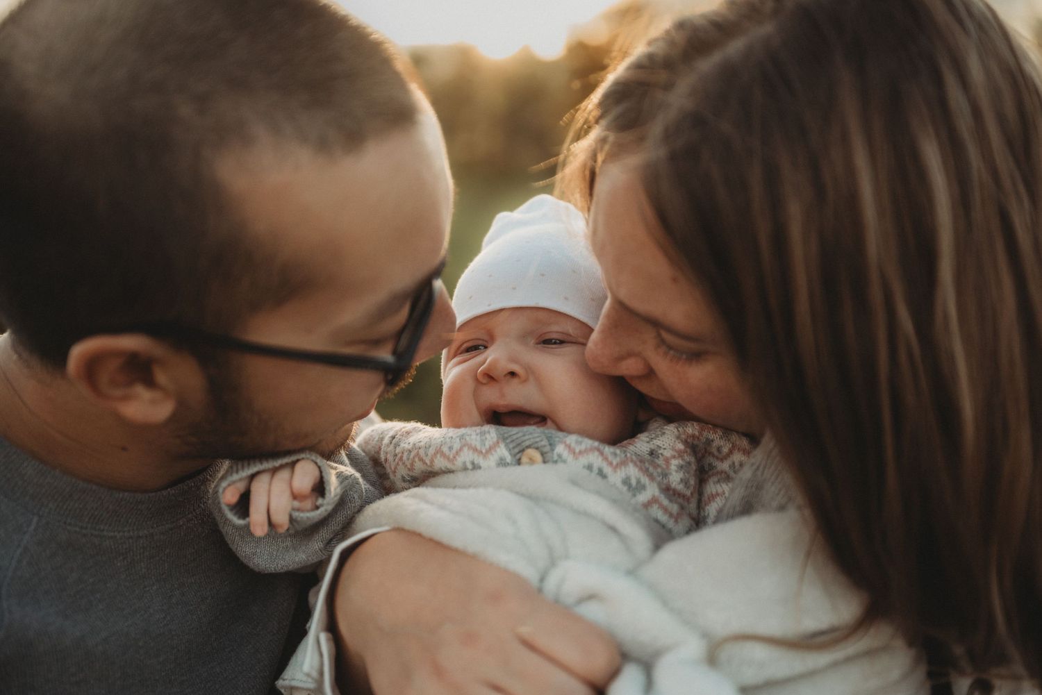 Parents kiss their laughing baby during outdoor family photoshoot.