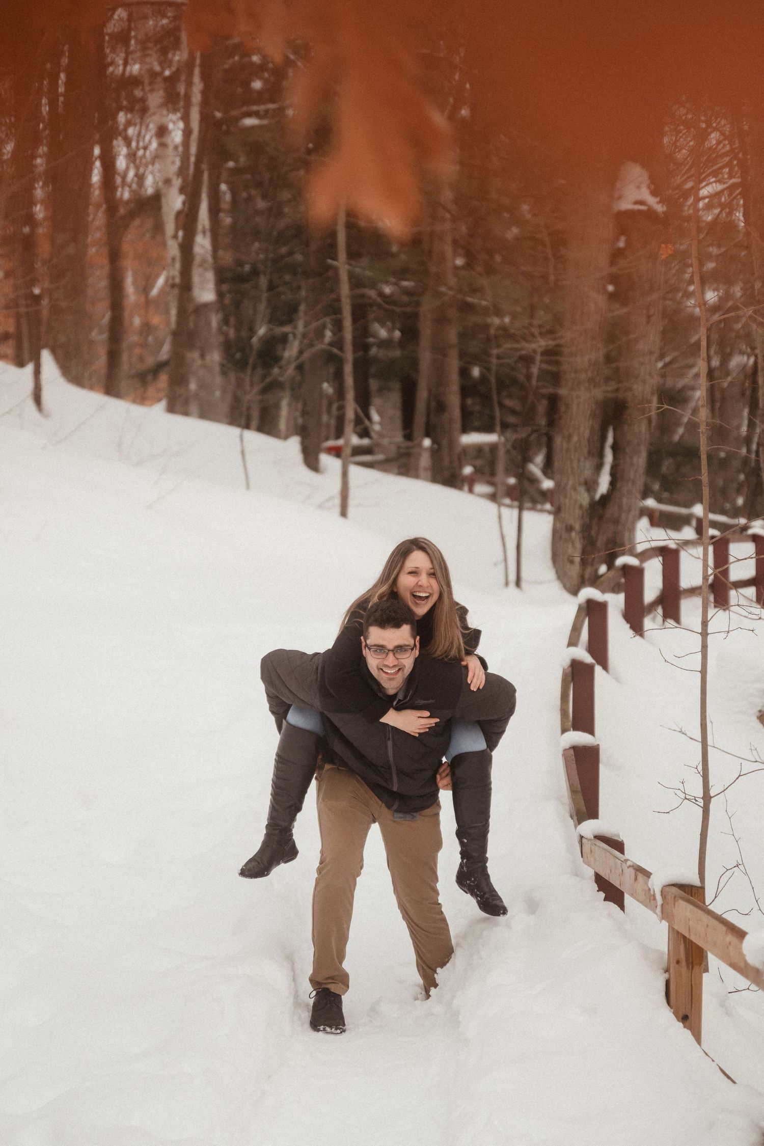 Couple enjoying a piggyback ride along a snowy forest path with warm sunset lighting.
