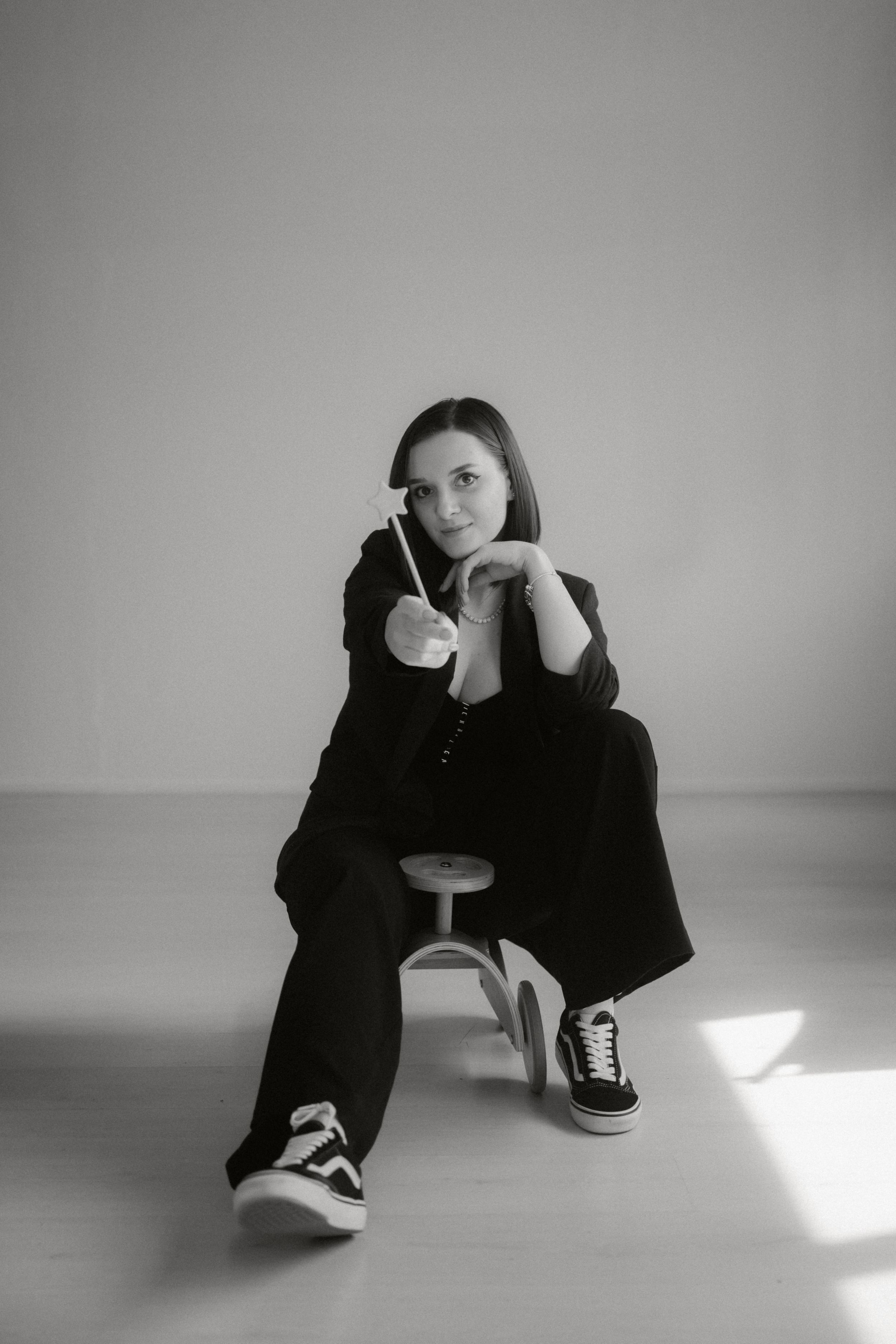 Black and white portrait of a person in dark attire and Vans sneakers sitting on a stool in a minimalist setting.