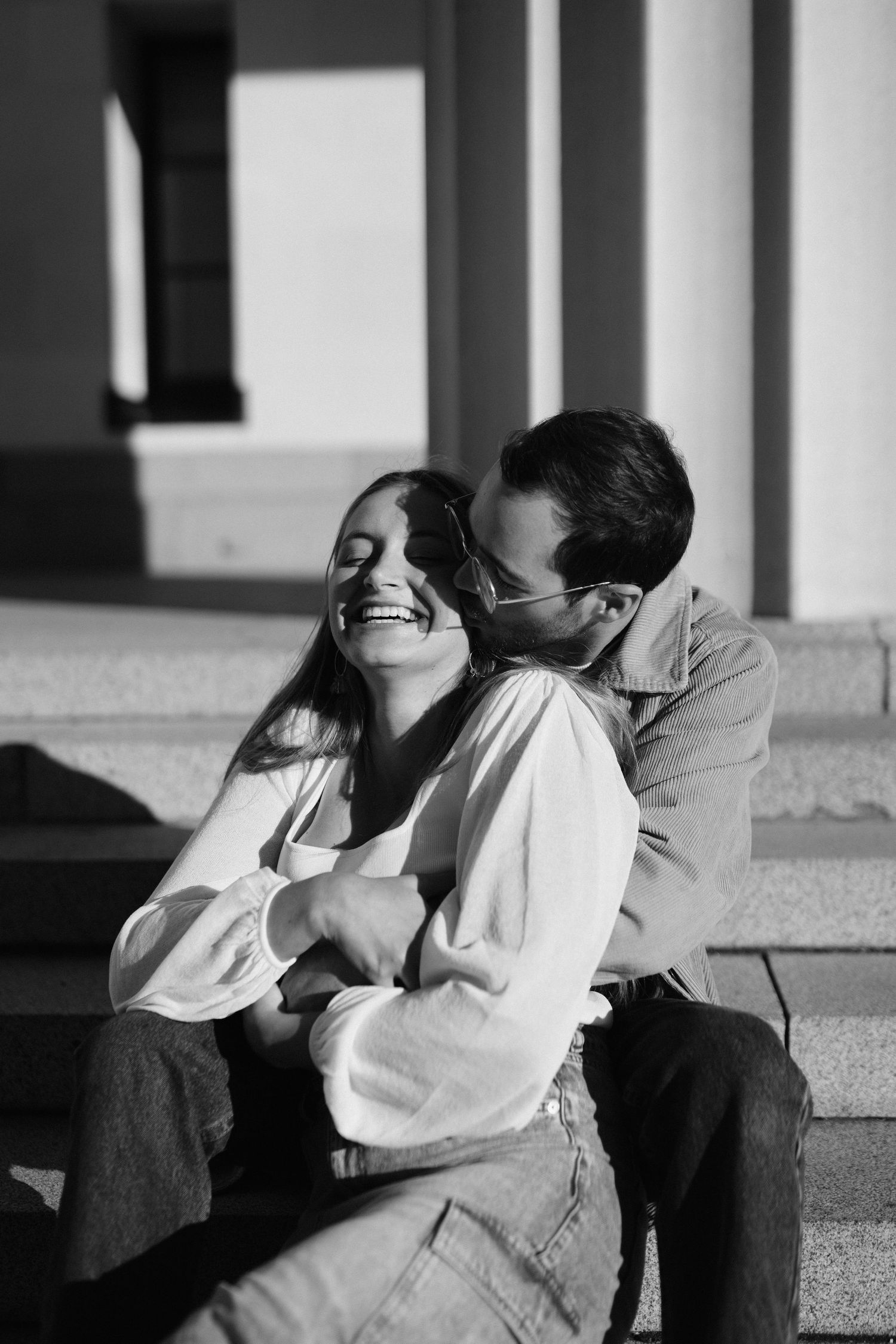 A black and white candid photo of a couple sharing a joyful embrace on stone steps.