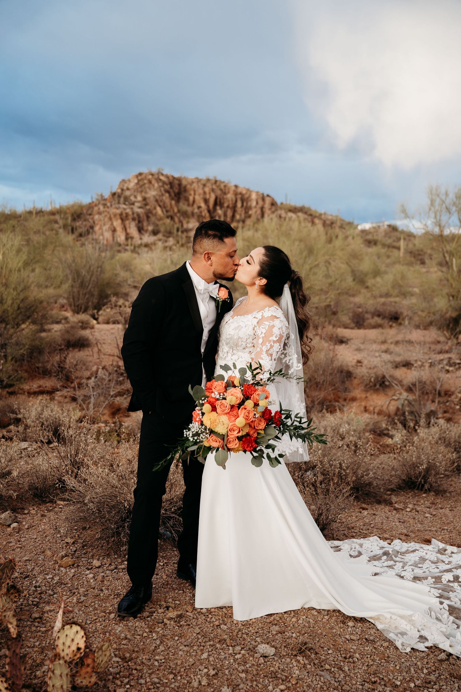 A couple shares a romantic moment in the desert with rocky mountains and desert brush in the background.